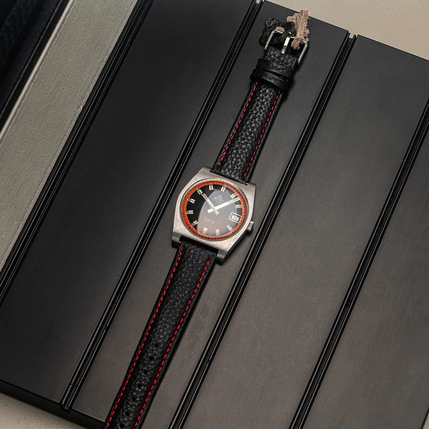 A wristwatch Rigi 2300-EC with a square silver-toned case, dark dial, and textured black leather strap with red stitching, displayed diagonally on a black presentation box.