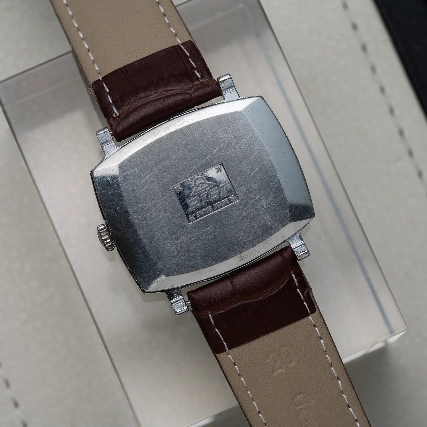 Close-up of the back of a vintage wristwatch Rigi 2300-EC, Cal. AS 1950 with a brushed silver-tone metal case and a brown leather strap, resting on a light-colored surface.