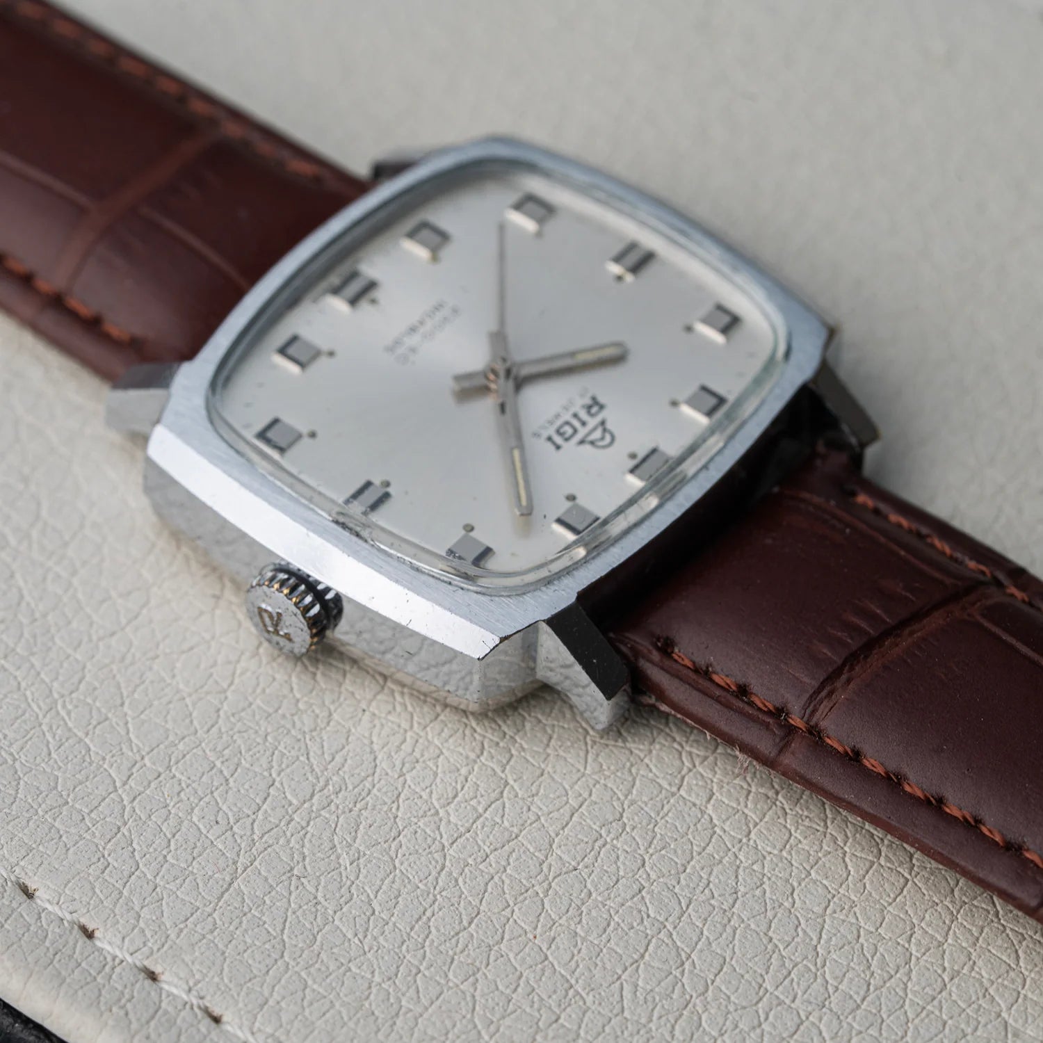 A vintage-style wristwatch with a silver-toned square case, silver dial, and dark brown leather strap, photographed close-up on a light textured surface.