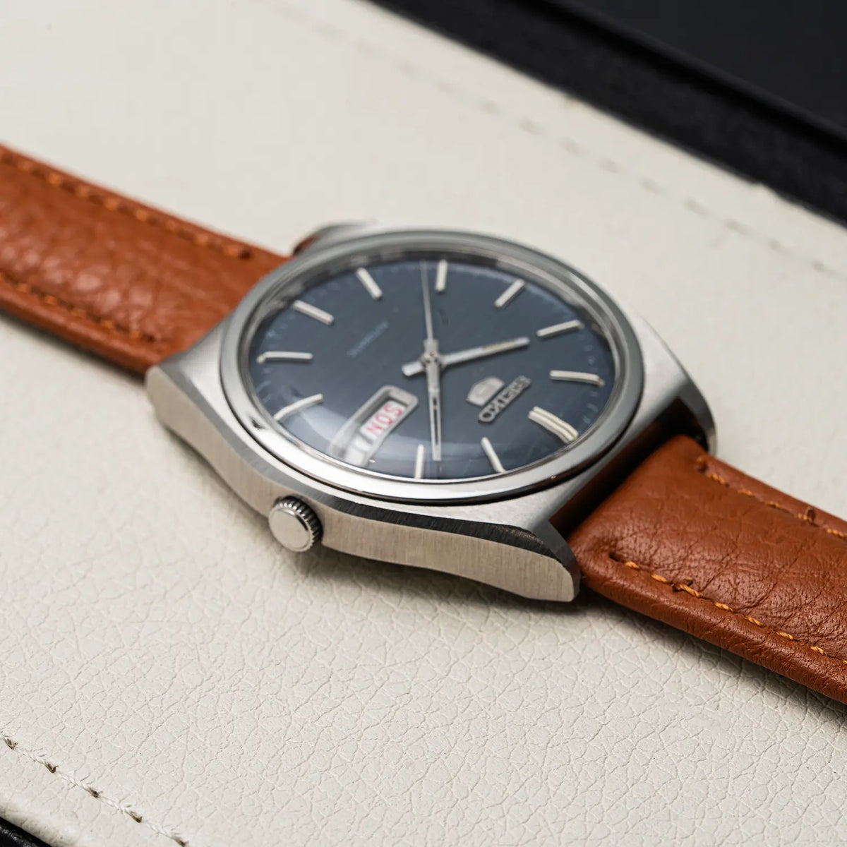 A close-up of an Seiko 5, Ref. 6319-8100, Cal. 6319, Men’s Automatic, 1977wristwatch with a stainless steel case, dark blue dial, and brown leather strap, resting on a textured off-white leather surface.