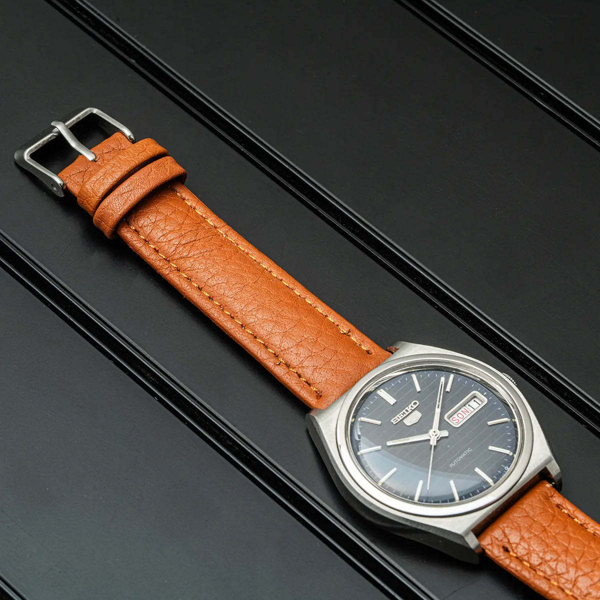 A Seiko 5, Ref. 6319-8100, Cal. 6319, Men’s Automatic, 1977 analog watch with a textured orange-brown leather strap and a silver-toned case, displayed diagonally on a dark textured background.