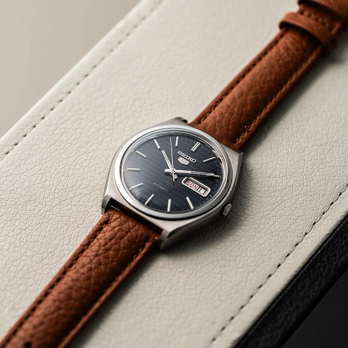 A Seiko 5, Ref. 6319-8100, Cal. 6319, Men’s Automatic, 1977 wristwatch with a dark blue sunburst dial, silver-toned case, and a brown textured leather strap, resting on a light beige leather surface.