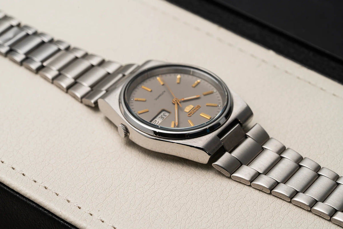 A vintage-style wristwatch Seiko 5, Ref. 7009-3130, Cal. 7009, Men’s Automatic, 1984 with a silver metal link bracelet, a round stainless steel case, and a dark gray dial with gold-tone hour markers and hands. The watch is resting on a textured off-white leather surface.