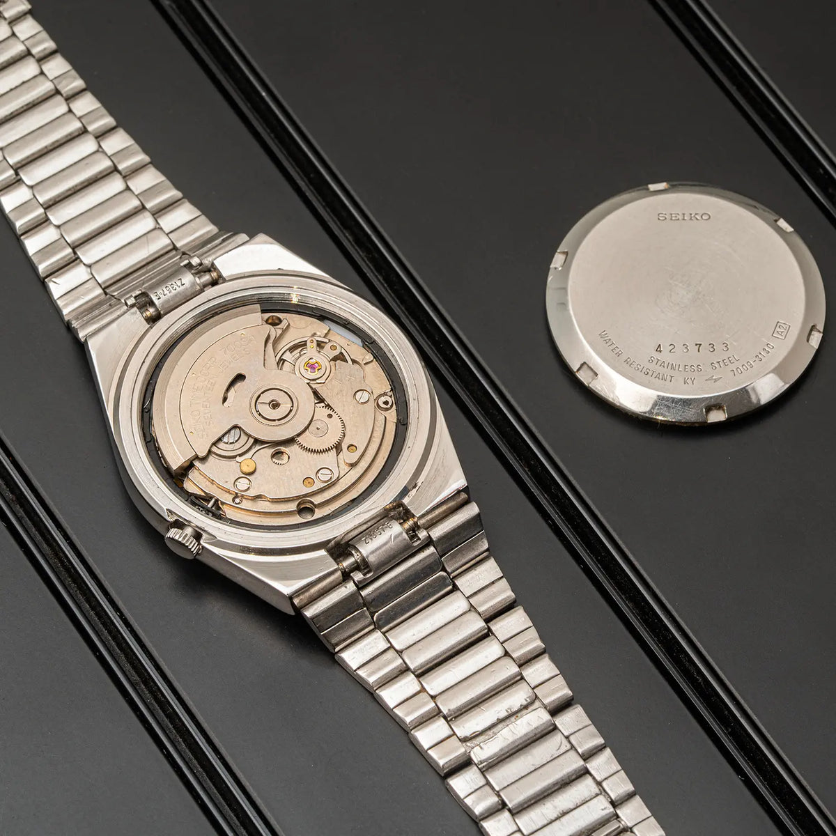 A silver stainless steel wristwatch Seiko 5, Ref. 7009-3130, Cal. 7009, Men’s Automatic, 1984 with a transparent case back showing the mechanical movement, displayed on a black surface next to its circular silver back cover.