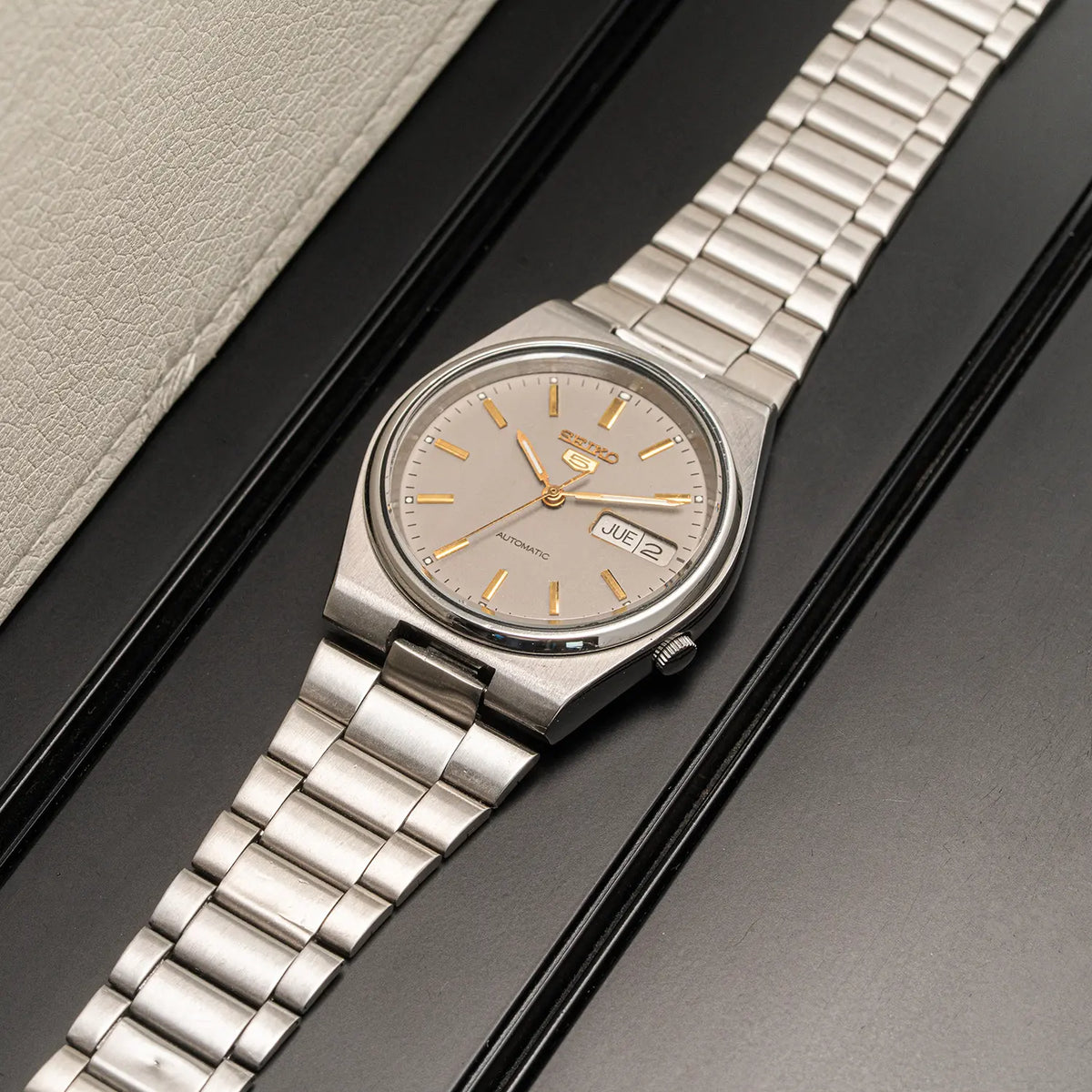 A vintage-style stainless steel wristwatch Seiko 5, Ref. 7009-3130, Cal. 7009, Men’s Automatic, 1984 with a silver metal link bracelet and a beige dial, photographed diagonally on a black surface next to a light beige textured surface.