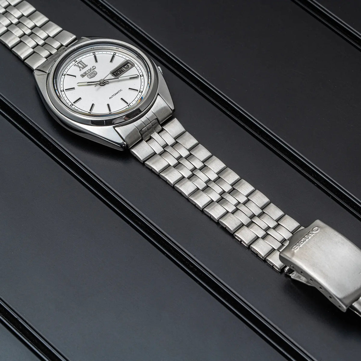 A silver stainless steel wristwatch Seiko 5, Ref. 7009-3140, Cal. 7009, Men’s Automatic, 1979 with a white dial and date window, resting diagonally on a dark textured background.