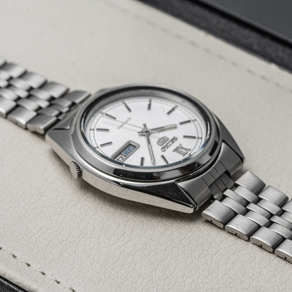 A close-up of a wristwatch Seiko 5, Ref. 7009-3140, Cal. 7009, Men’s Automatic, 1979 with a silver-toned metal bracelet and a white dial, resting on a light textured surface.