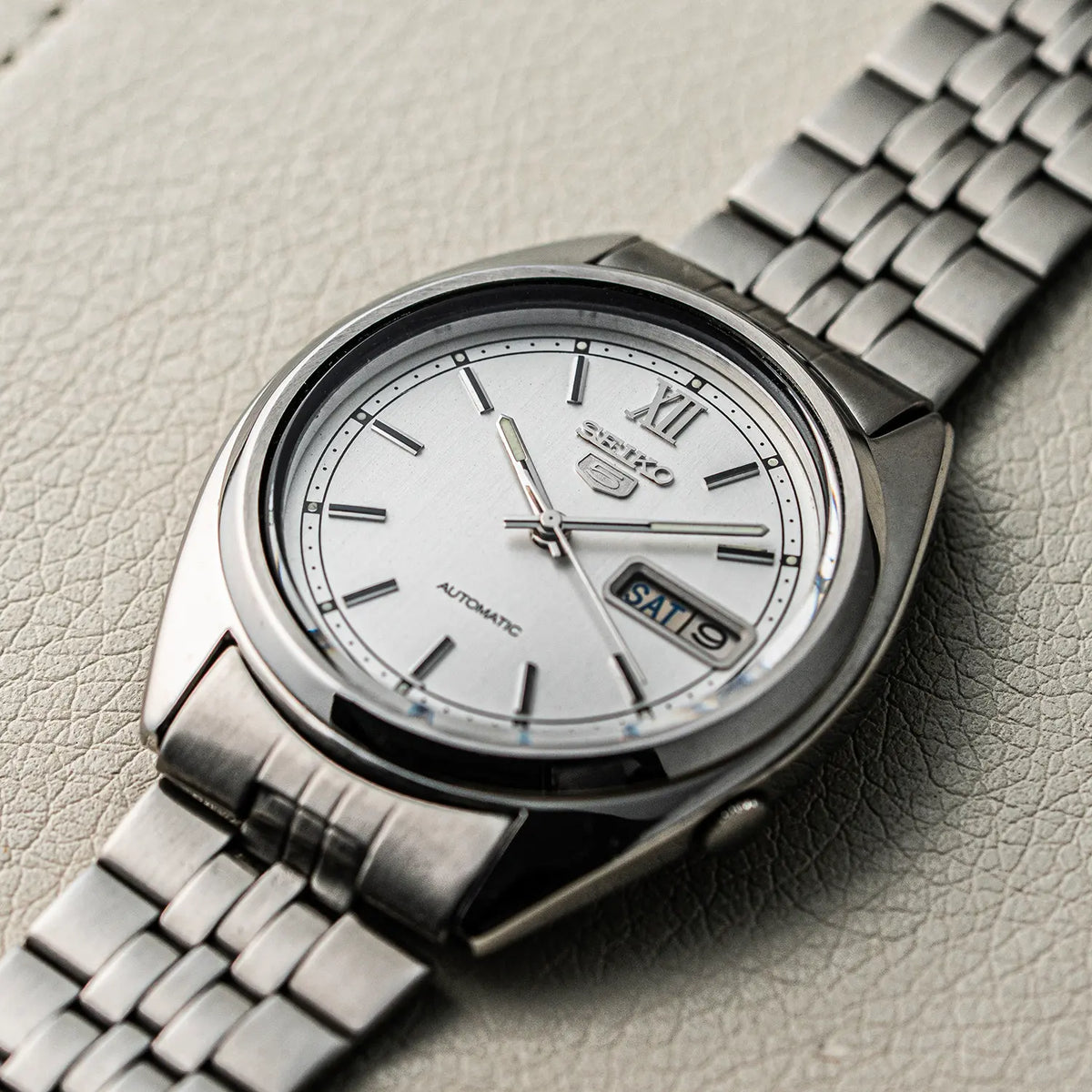 A Seiko 5, Ref. 7009-3140, Cal. 7009, Men’s Automatic, 1979 automatic wristwatch with a silver-toned metal bracelet and a white dial, photographed diagonally on a textured light gray surface.