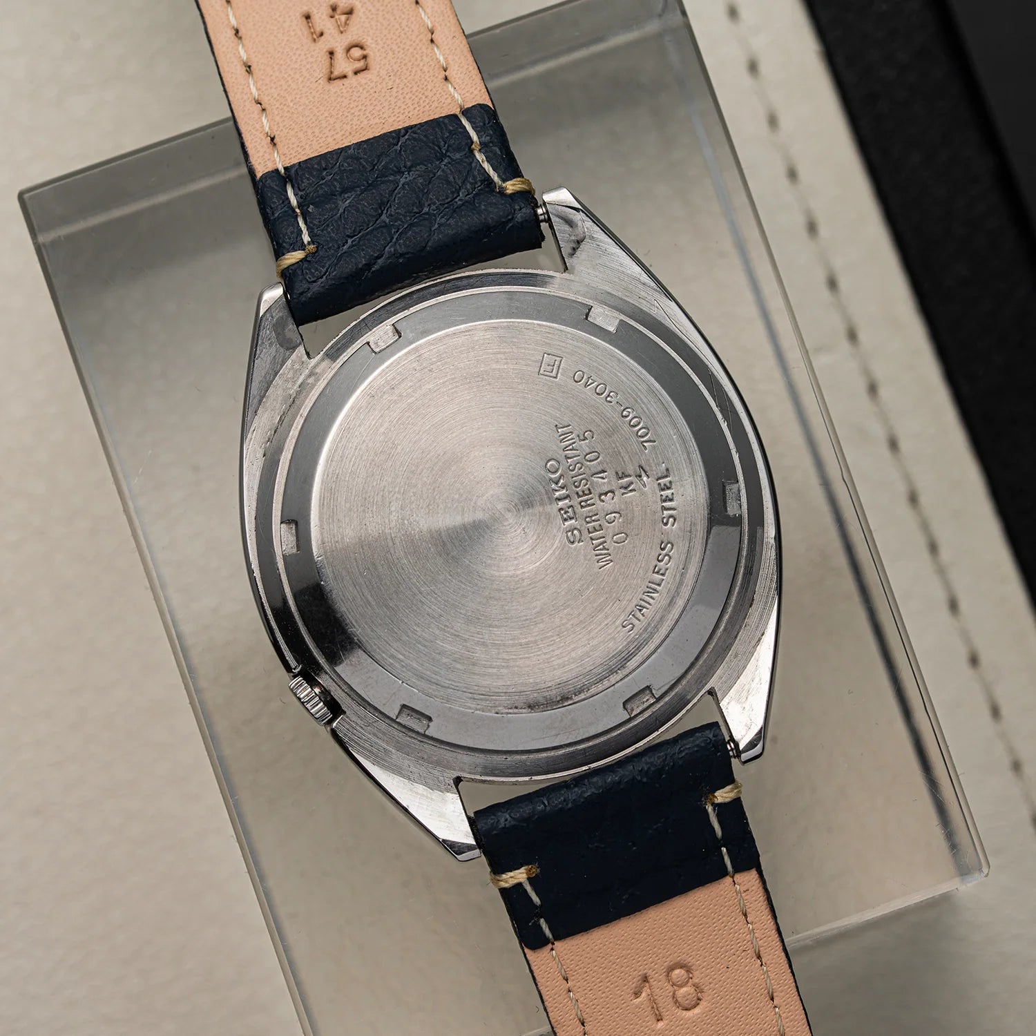 Close-up of the back of a vintage wristwatch Seiko 5 7009-3040 with a stainless steel case and a tan leather strap, resting on a clear display box.
