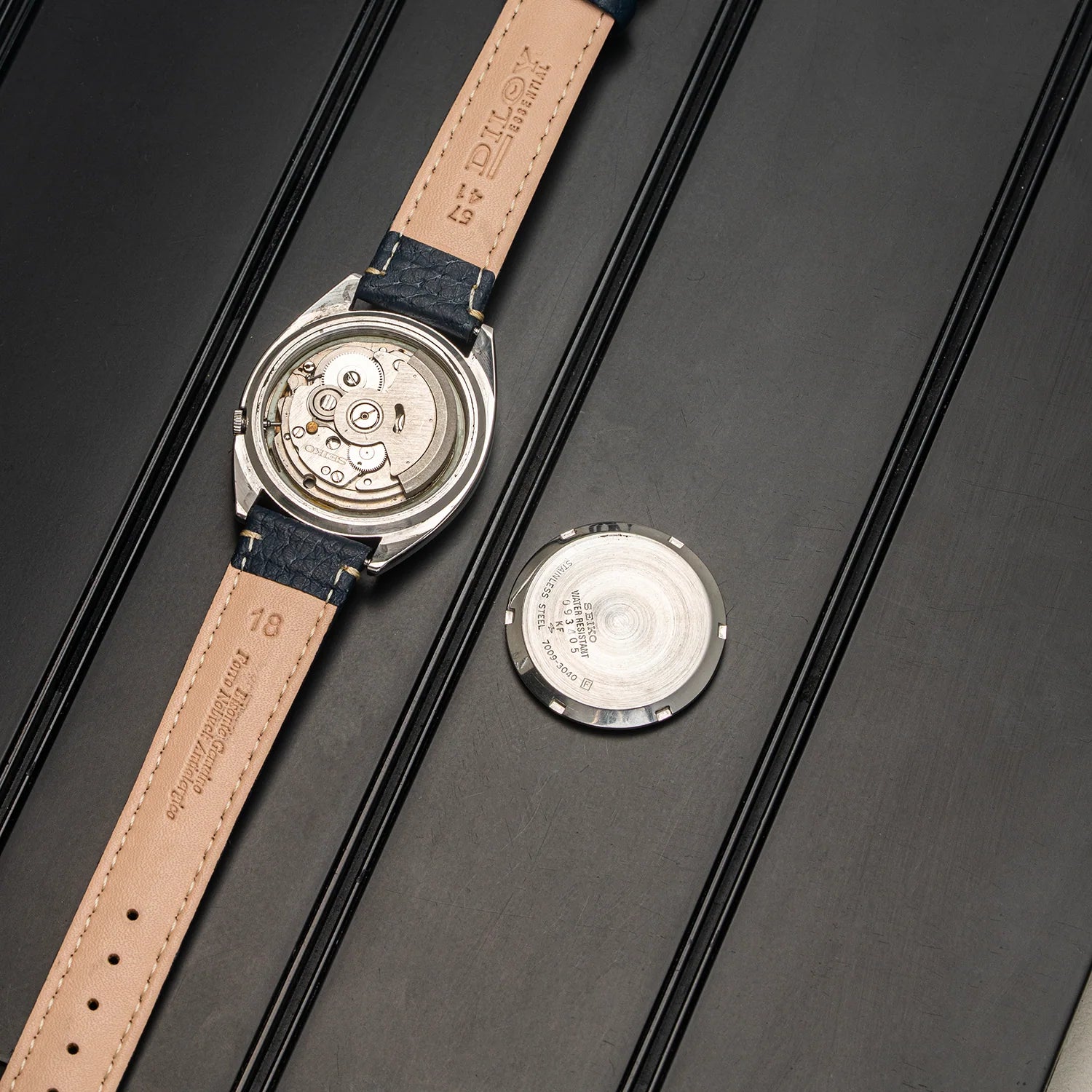 A luxury watch shown from the back, featuring a beige leather strap with debossed branding and a dark blue fabric section, alongside its separate silver metal case lid on a dark textured background.