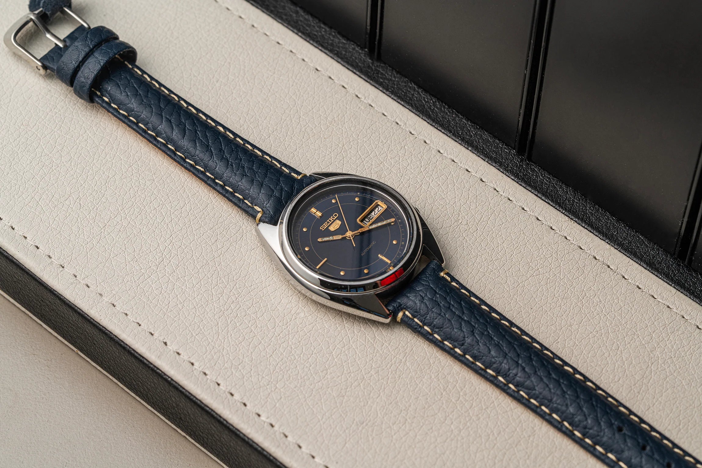 A vintage-style wristwatch Seiko 5 7009-3040 with a dark navy blue leather strap, a silver-toned case, and a matching dark dial, resting diagonally on a cream-colored textured leather surface.