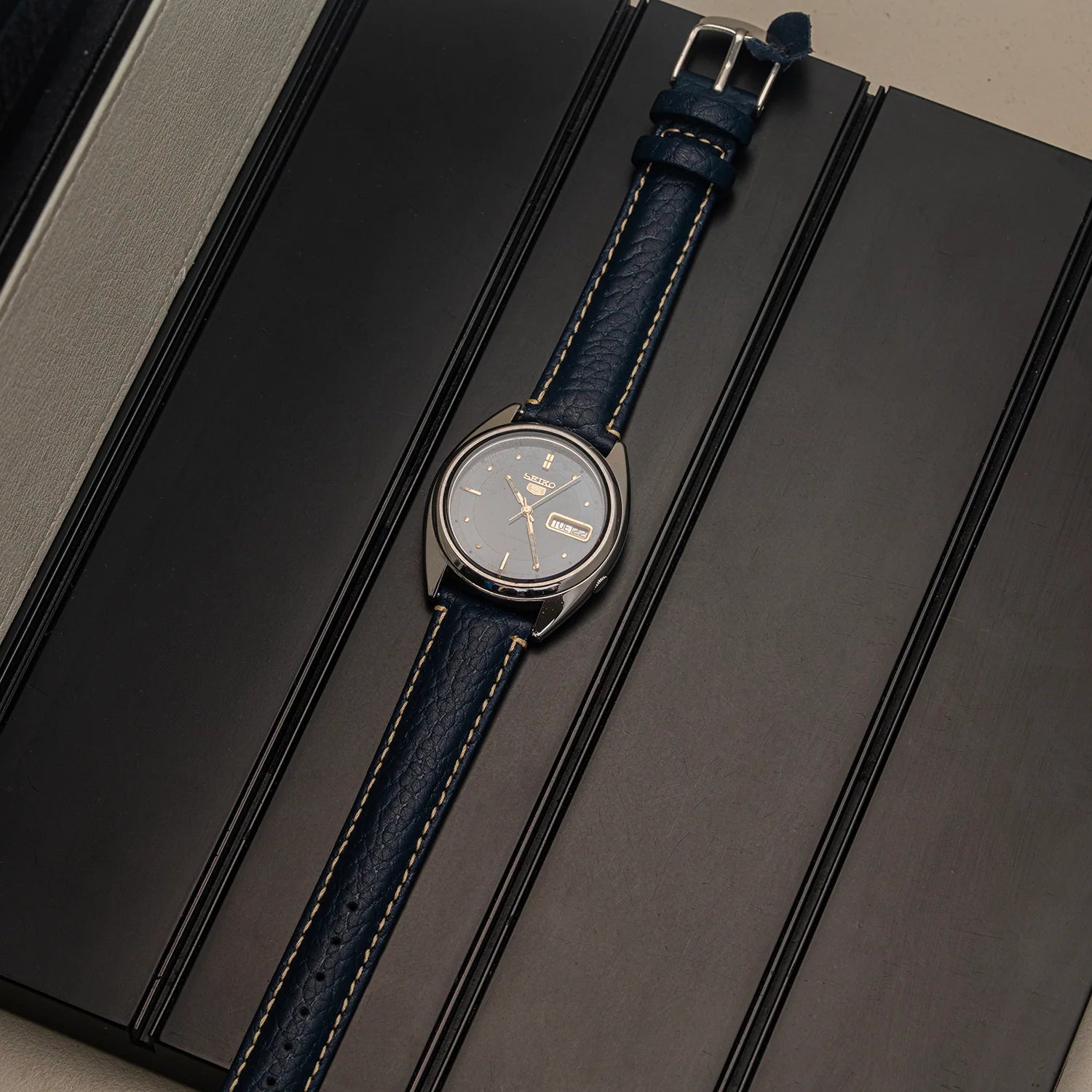 A wristwatch with a dark blue textured leather strap and a silver-toned round case, displayed diagonally on a dark gray presentation box.