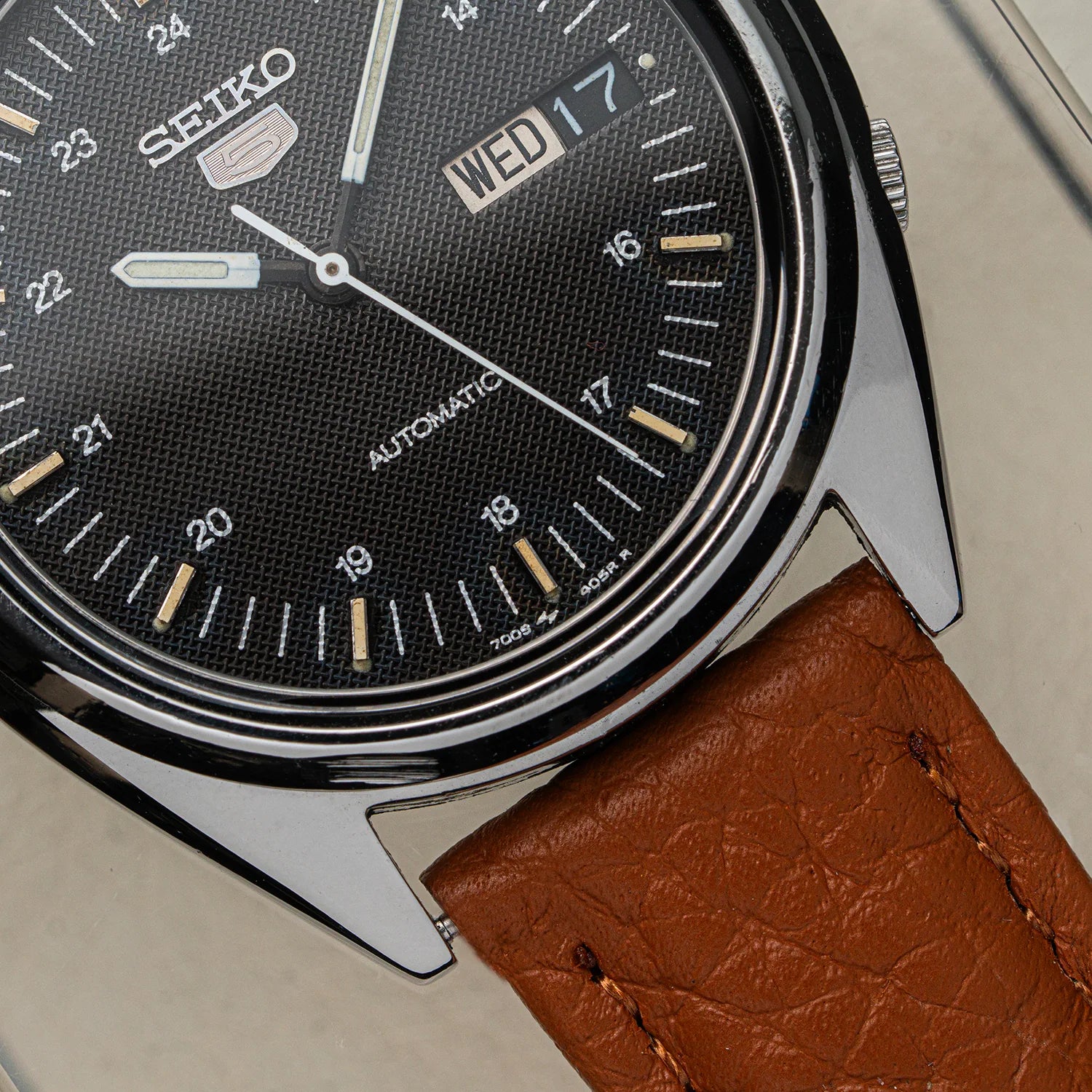 Close-up of a Seiko 5 7009-3040 watch with a black dial, silver-tone case, and brown leather strap on a light neutral background.