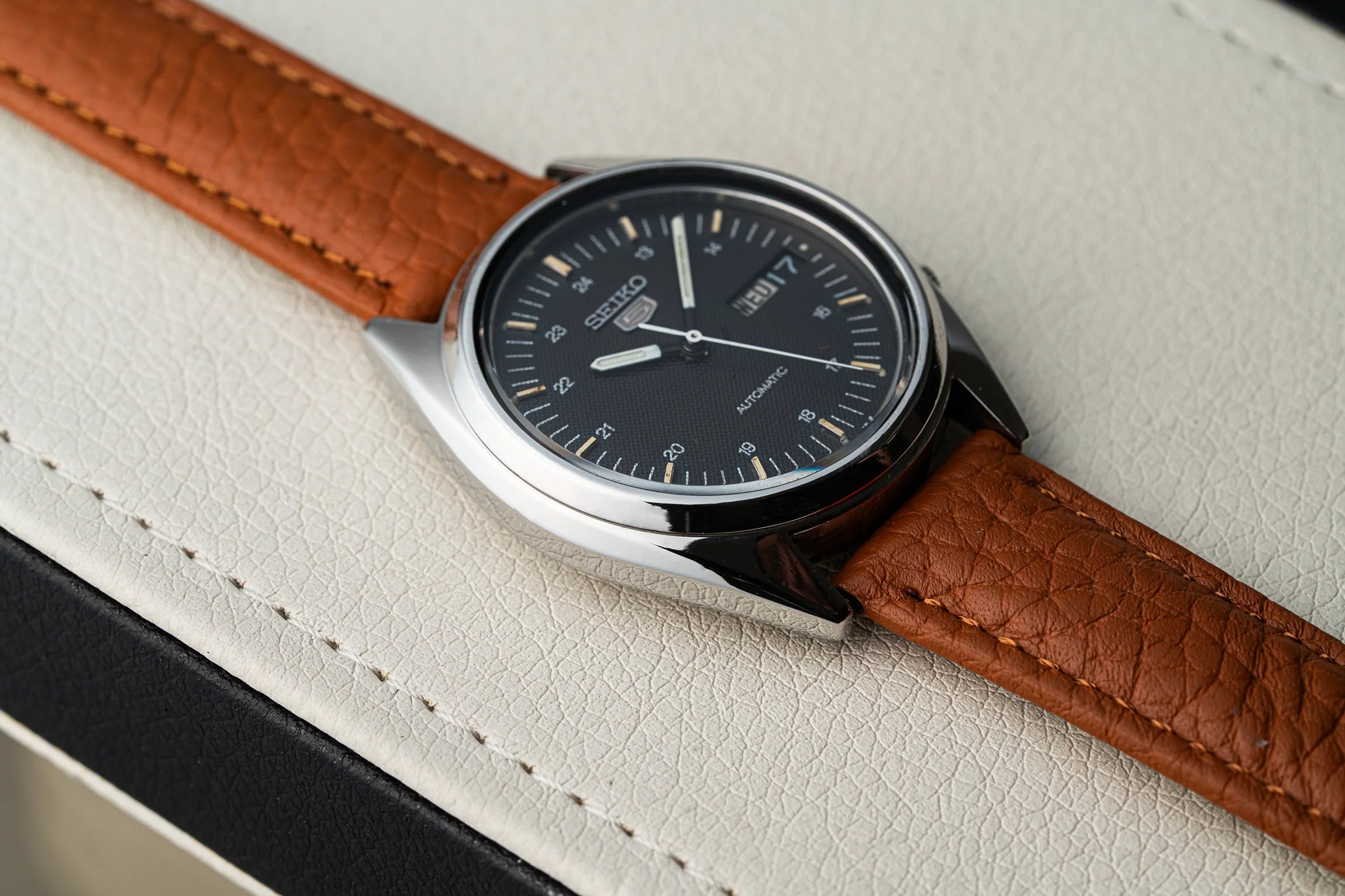 A close-up shot of a wristwatch Seiko 5 7009-3040 with a brown textured leather strap and a black dial, resting on a white textured leather surface.