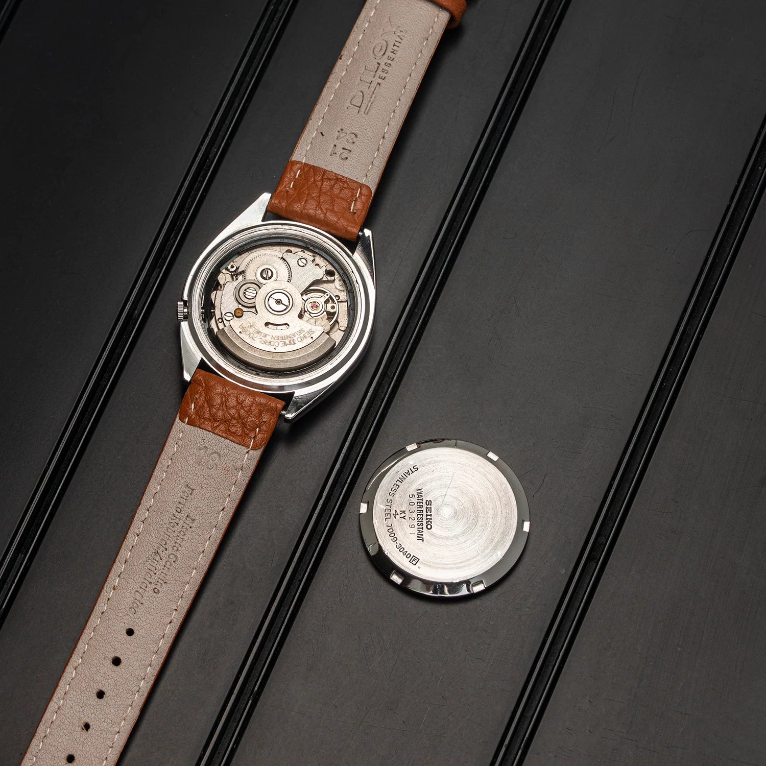 A luxury watch Seiko 5 7009-3040 with a silver-toned case, featuring a light beige leather strap with brown alligator-textured accents, shown with its back casing removed and placed beside it on a dark textured background.