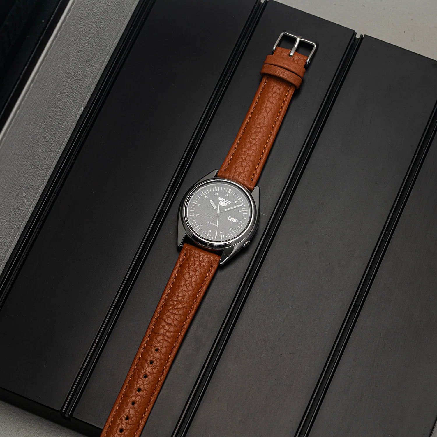 A wristwatch Seiko 5 7009-3040 with a brown textured leather strap and a silver-toned oval dial, laid diagonally on a black segmented surface.