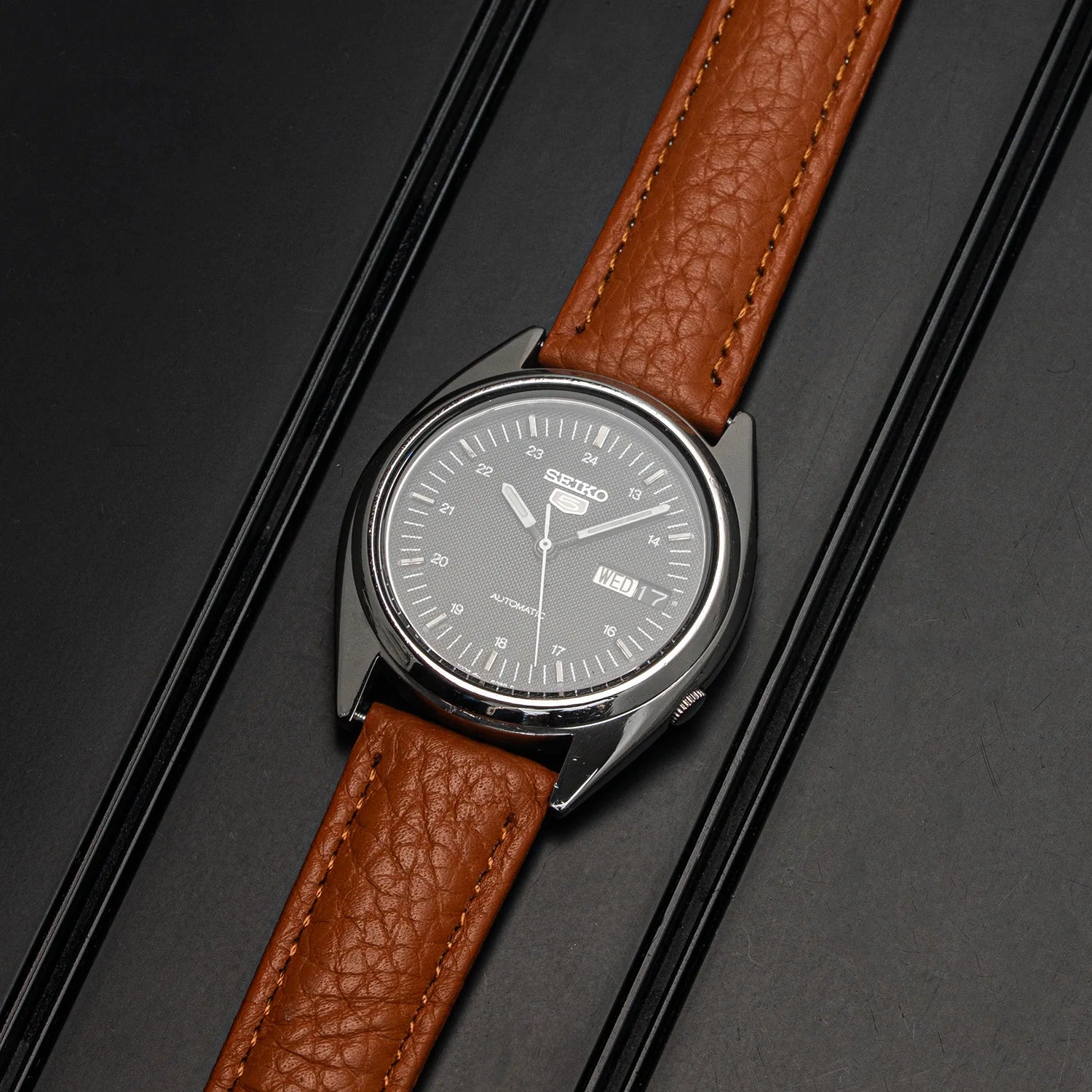 A Seiko wristwatch Seiko 5 7009-3040 with a silver-toned oval case, a light gray dial, and a textured brown leather strap, photographed diagonally on a dark textured background.