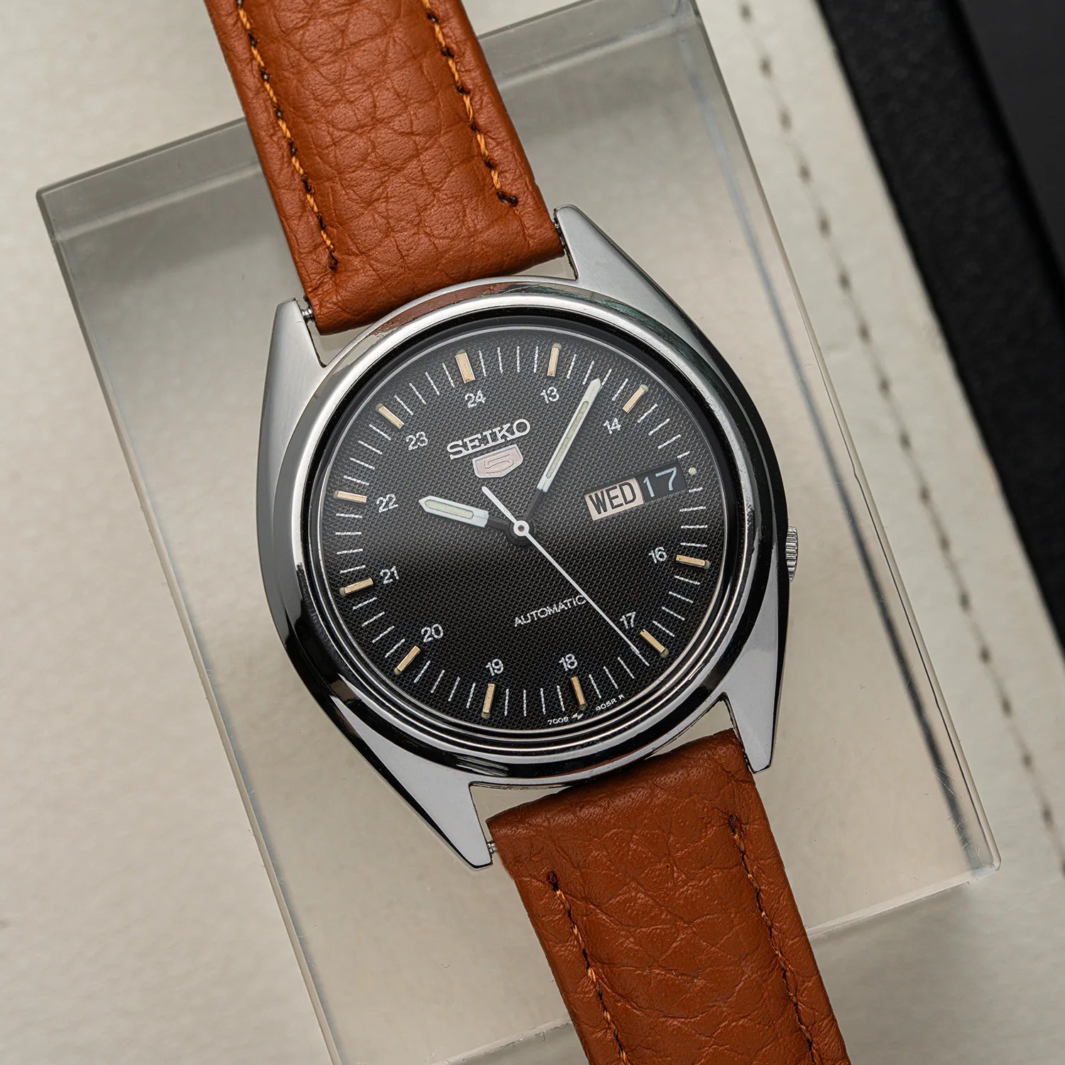 A Seiko automatic wristwatch Seiko 5 7009-3040 with a round silver-tone case, black dial, and brown leather strap, displayed on a clear acrylic stand against a light neutral background.
