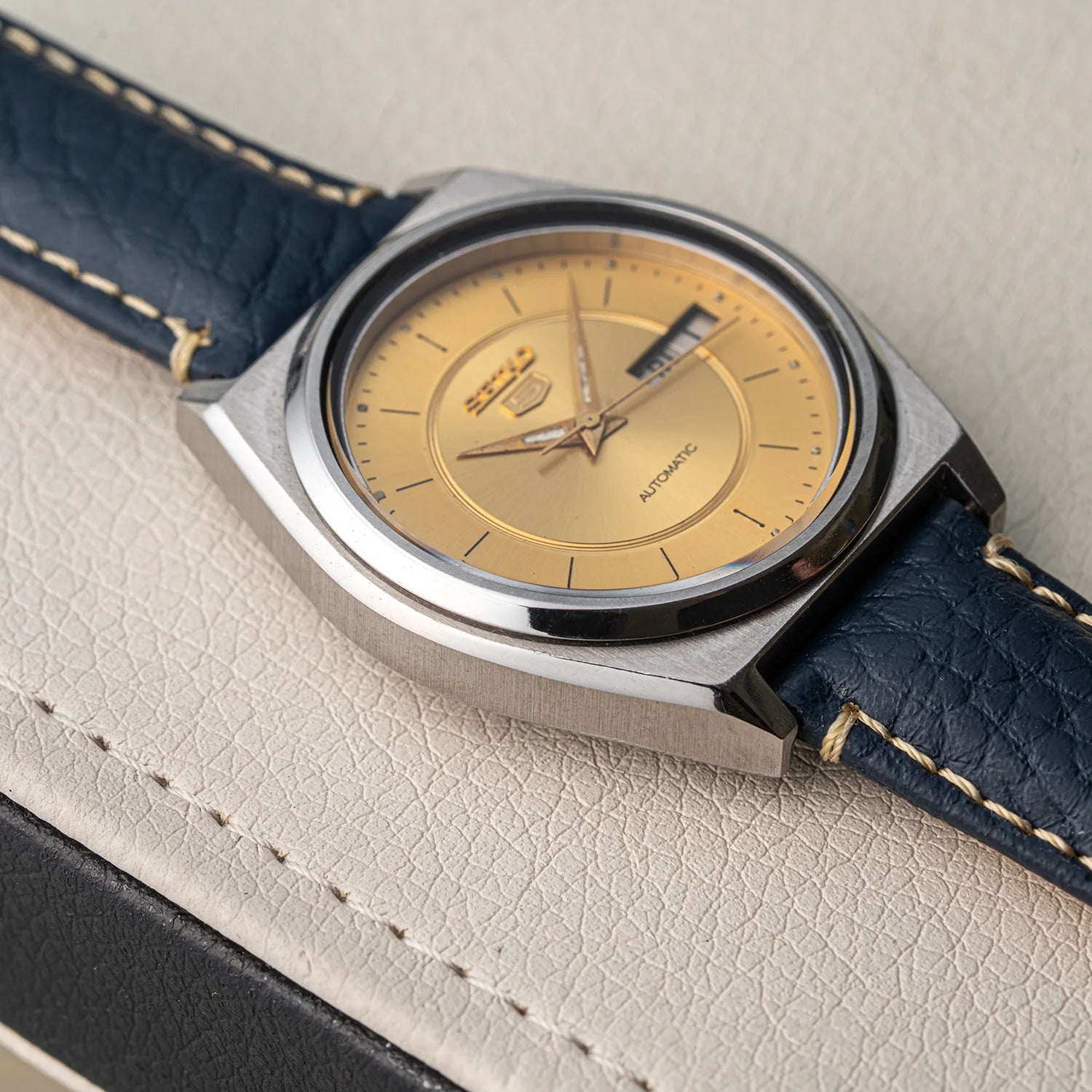 A vintage Seiko 5 7009-876A wristwatch with a gold dial and a dark blue leather strap, resting on a textured light beige leather surface.