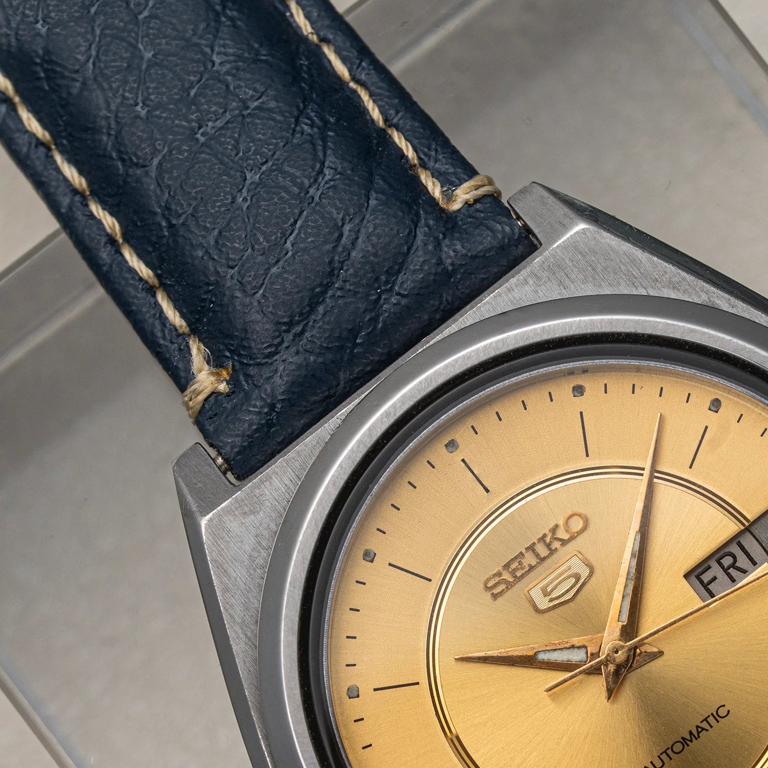 Close-up of a Seiko wristwatch Seiko 5 7009-876A with a brushed silver-tone case, gold dial, and dark blue leather strap with light tan stitching.
