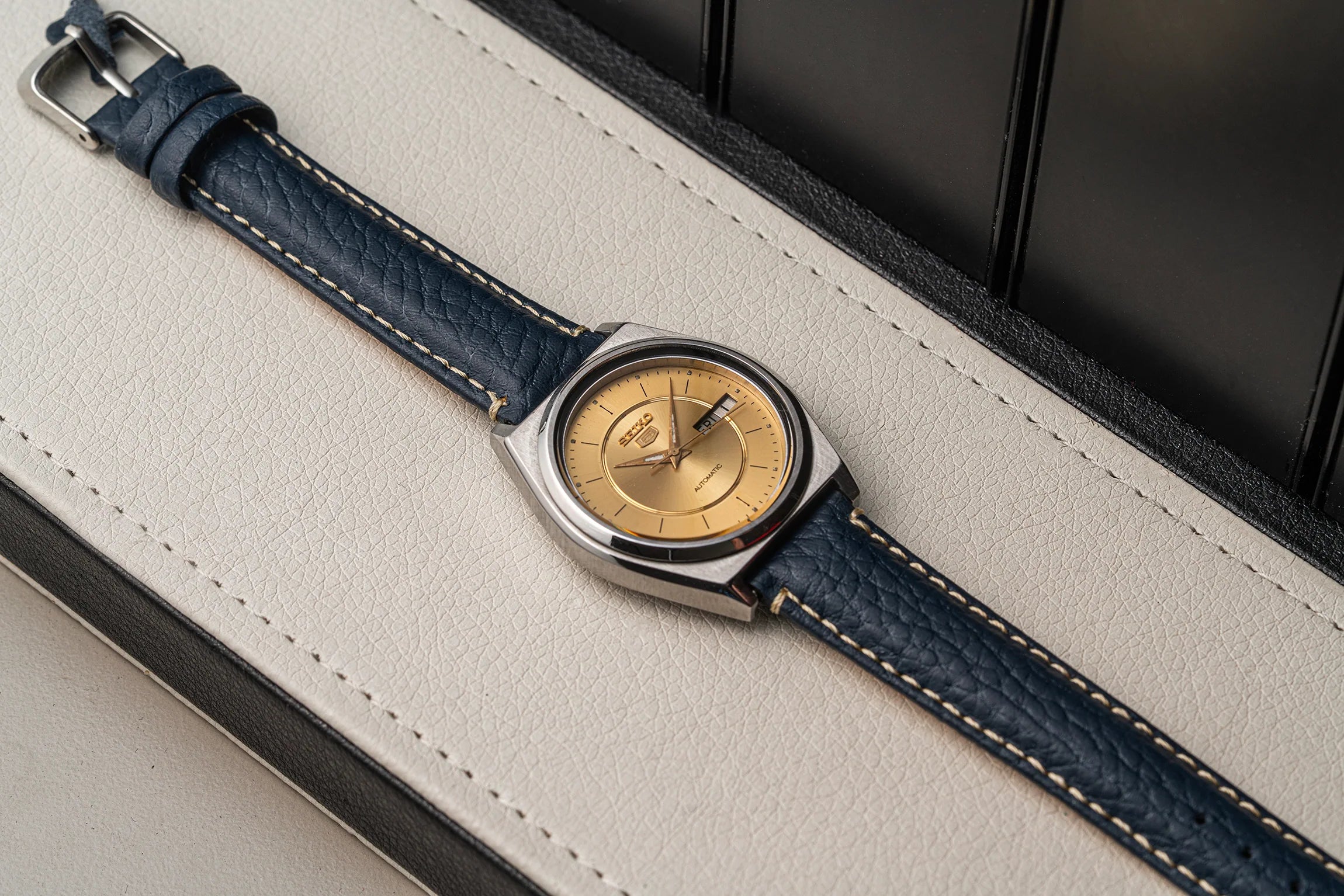 A wristwatch Seiko 5 7009-876A with a textured navy blue leather strap and a round gold dial, resting diagonally on a cream-colored textured surface.