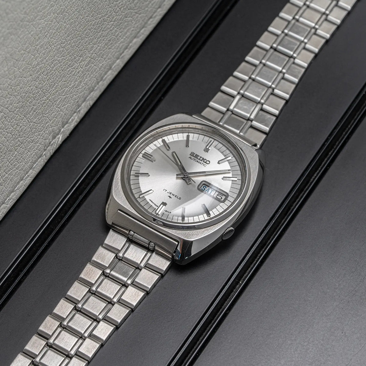 A Seiko, Ref. 7009-8100, Cal. 7009, Men’s Automatic, 1977 stainless steel watch with a silver dial and matching metal link bracelet, photographed diagonally on a dark textured surface next to a light gray textured insert.