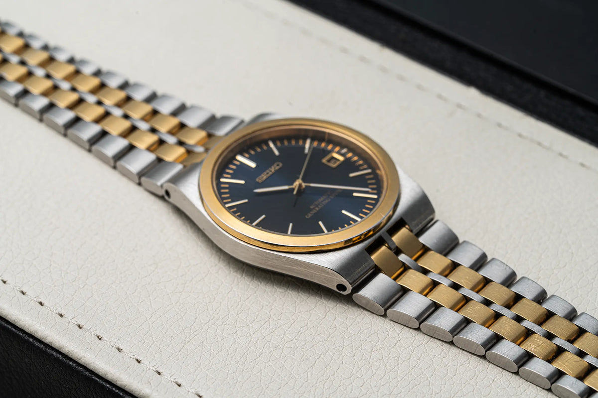 A close-up of a Seiko AGS, Ref. 5M22-8A10, Cal. 5M22, Men’s Quartz Analog watch with a two-tone silver and gold metal bracelet, a gold bezel, and a dark blue dial, resting on a textured off-white leather surface.