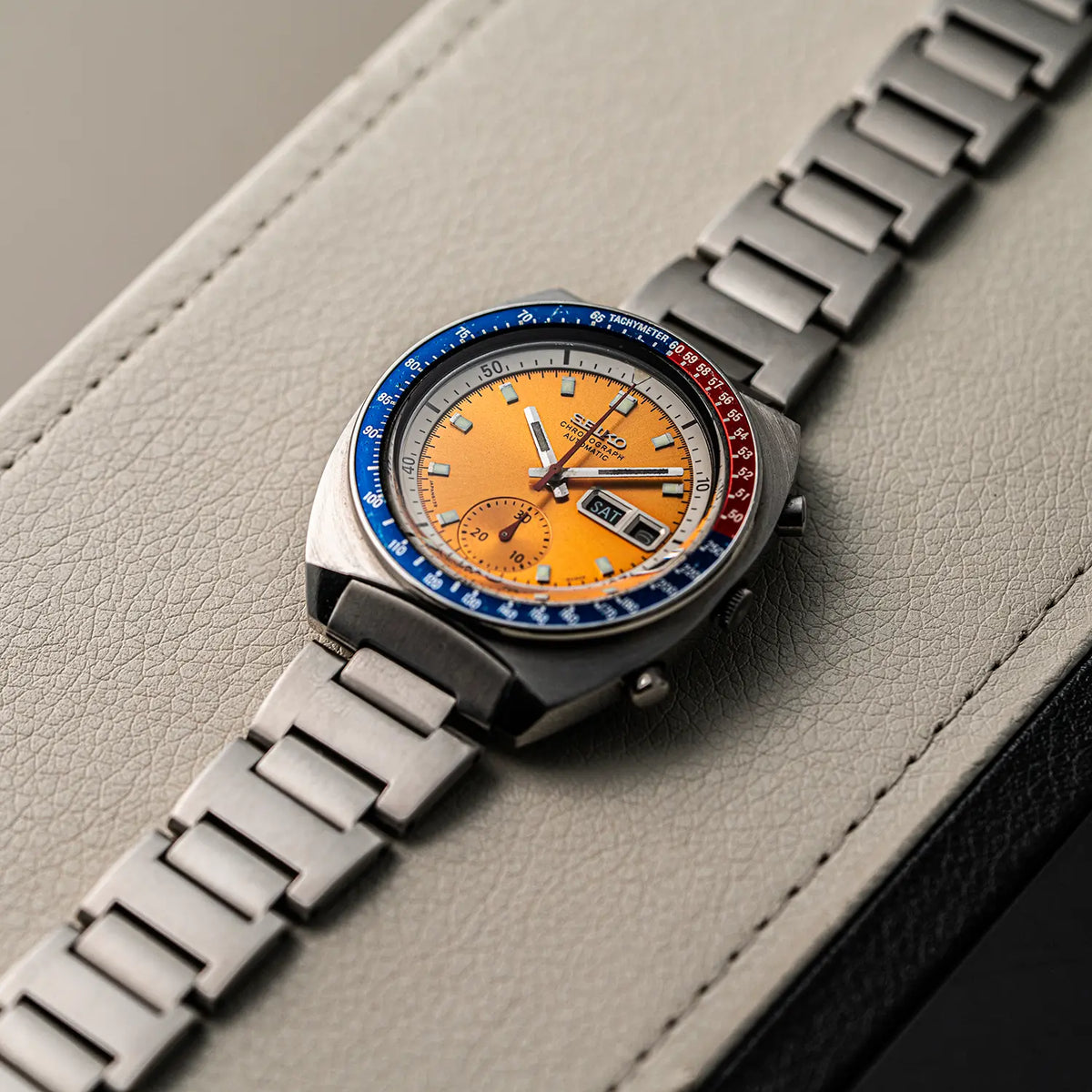 A wristwatch Seiko Chronograph Pogue, Ref. 6139-6002, Cal. 6139, Men’s Chronograph with a stainless steel link bracelet and a round orange dial, resting on a light gray textured leather surface. The bezel is split between blue and red sections with white markings.