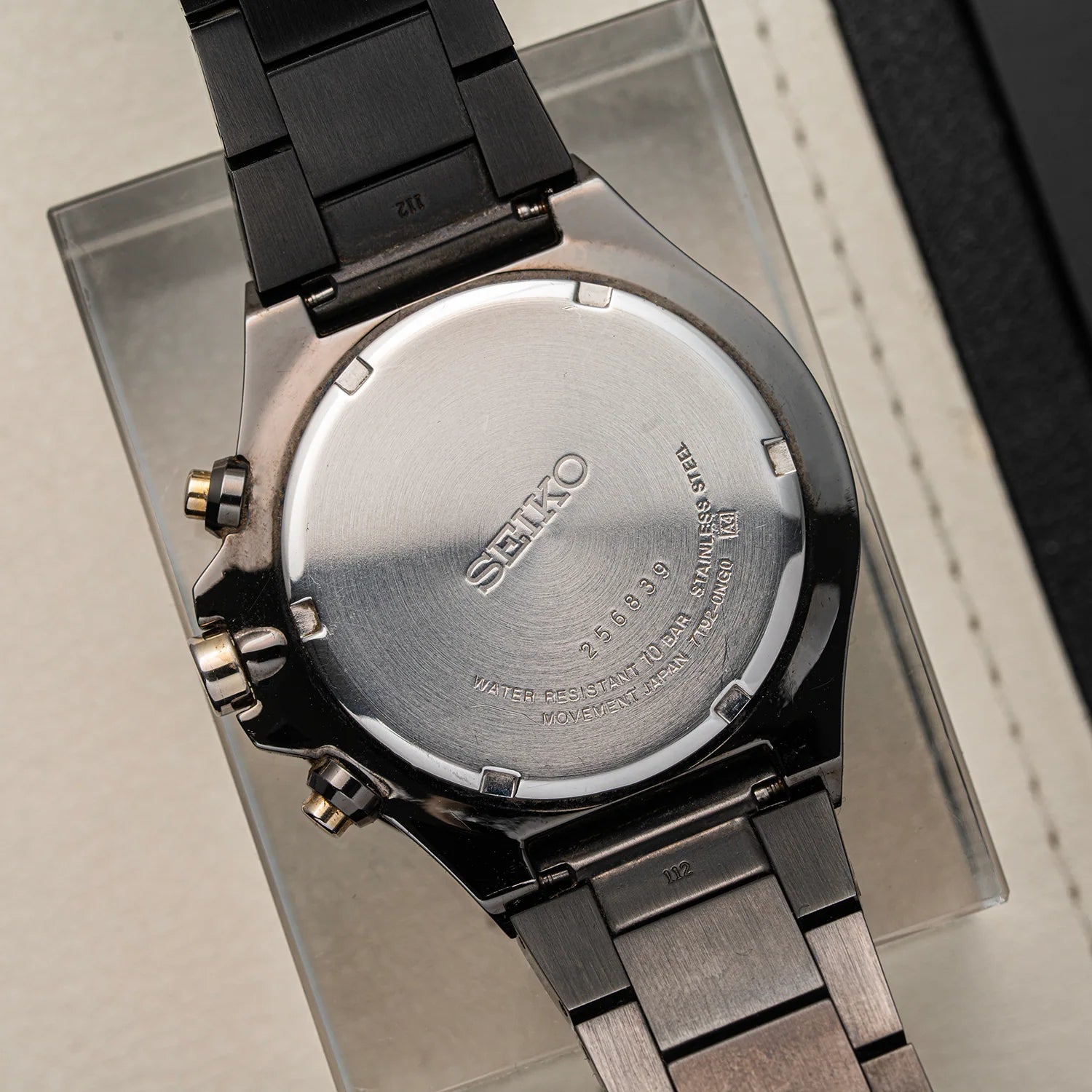 Close-up of the back of a Seiko Chronograph, Ref. 7T92-0NGO watch with a brushed stainless steel case and dark metal link bracelet, resting on a clear display stand.