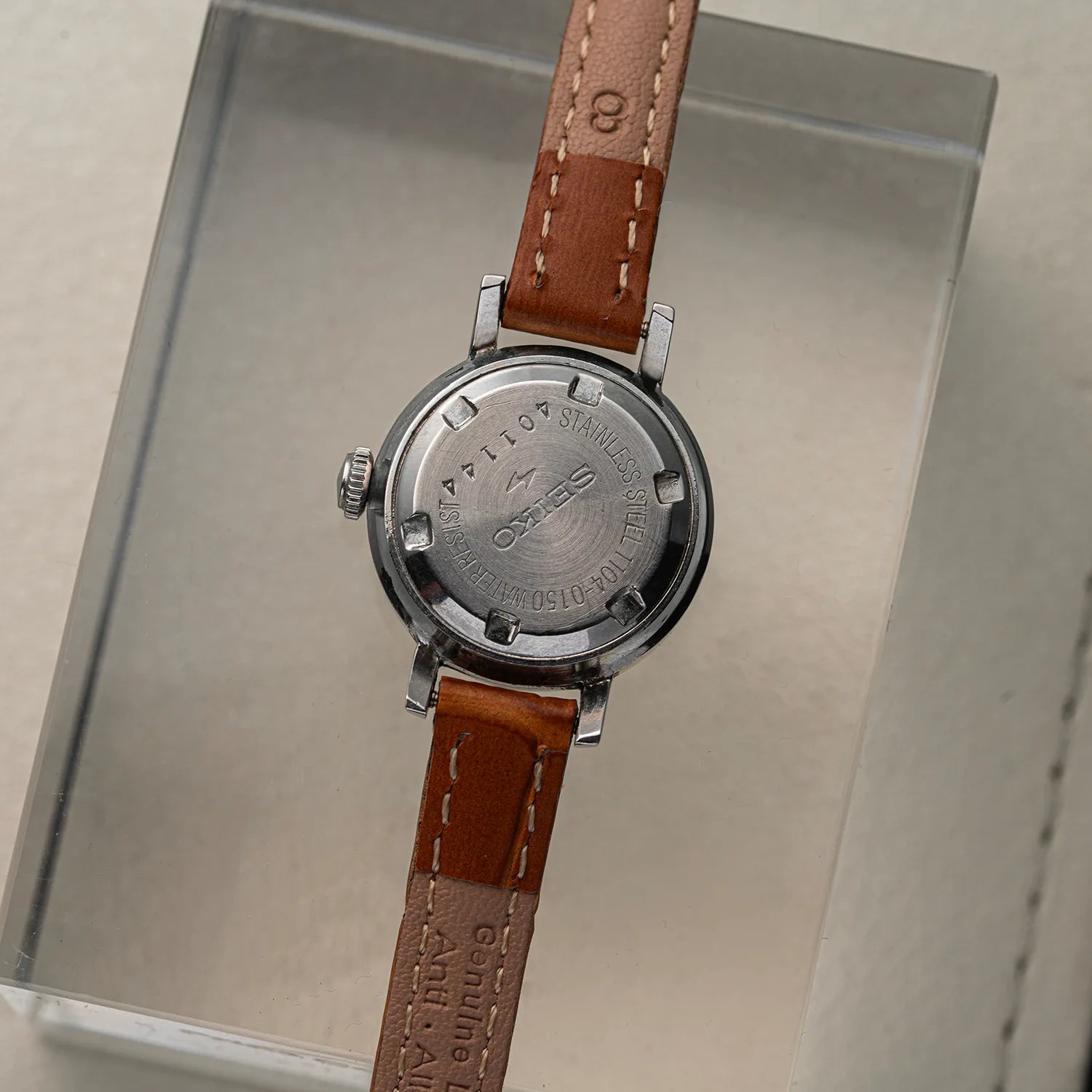 The back of an women's Seiko 1104-0150 watch with a brown leather strap and a stainless steel case, resting on a clear acrylic display block.
