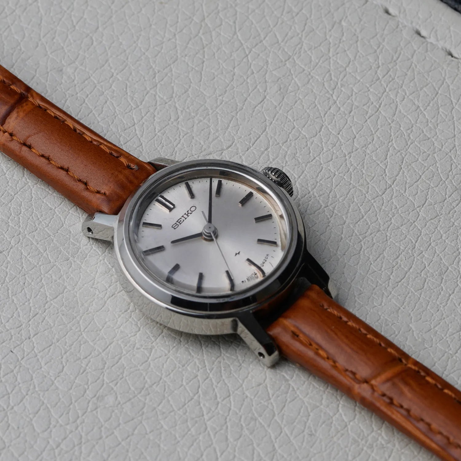 A women's Seiko 1104-0150 wristwatch with a silver-toned case, silver dial, and brown leather strap, photographed diagonally on a textured light gray surface.