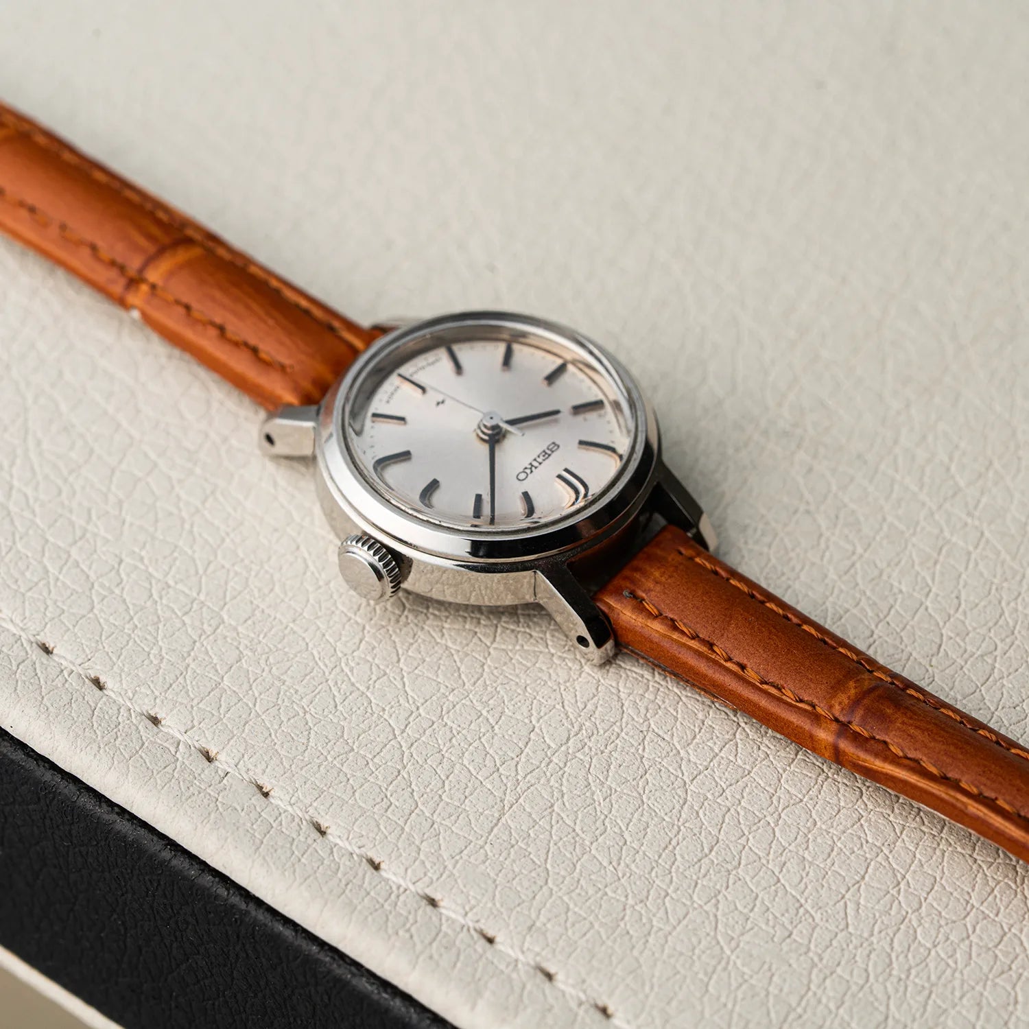 A close-up of a wristwatch women's Seiko 1104-0150 with a silver-toned case, white dial, and brown leather strap, resting on a textured off-white surface.