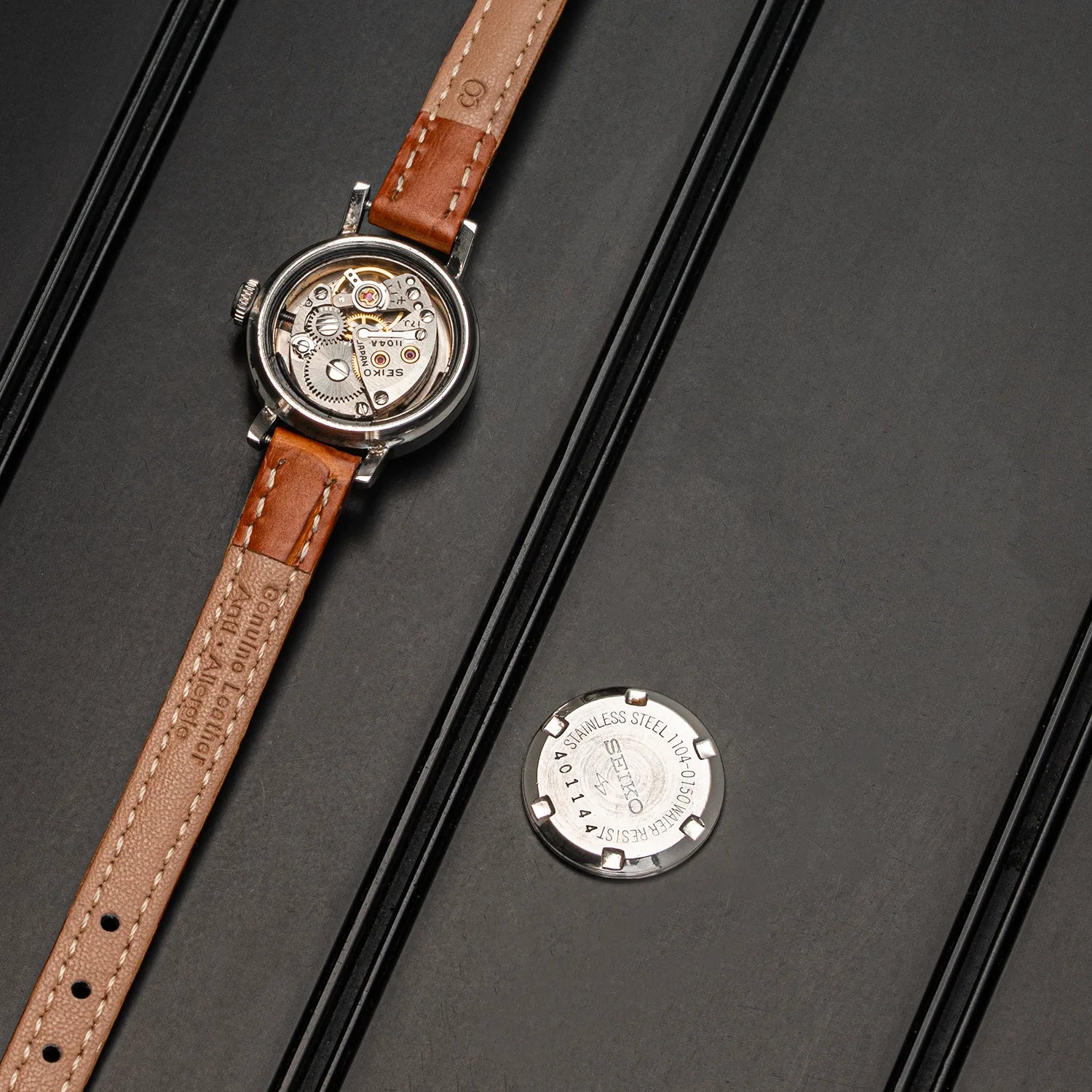 A luxury women's Seiko 1104-0150 watch with a brown leather strap and a round silver case, displayed diagonally on a dark textured background next to its circular silver backplate.