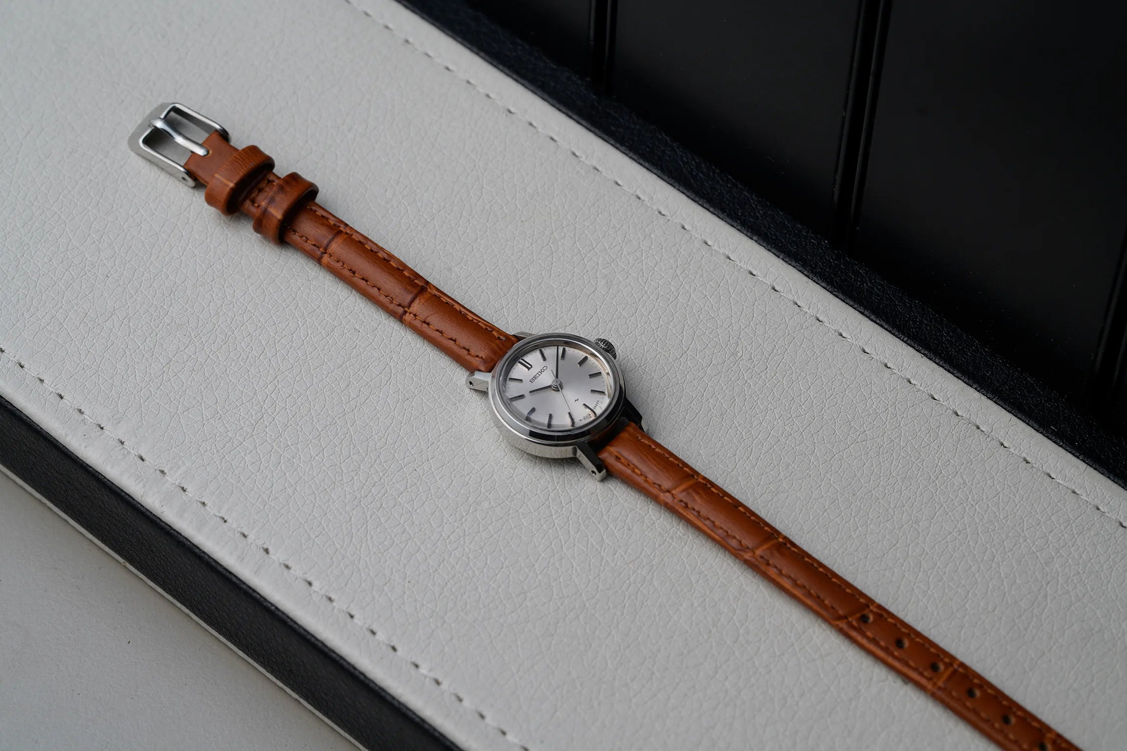 A vintage-style women's Seiko 1104-0150 wristwatch with a brown leather strap and a silver-toned round case, resting diagonally on a white textured leather surface with a black background.