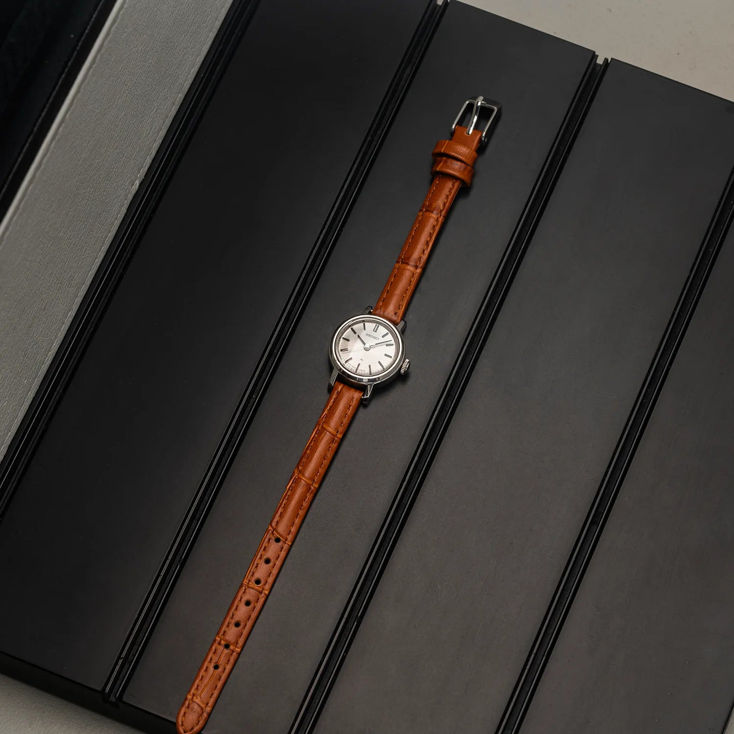 A vintage-style women's Seiko 1104-0150 wristwatch with a brown crocodile-embossed leather strap and a silver-toned round case, displayed diagonally on a dark gray textured background.