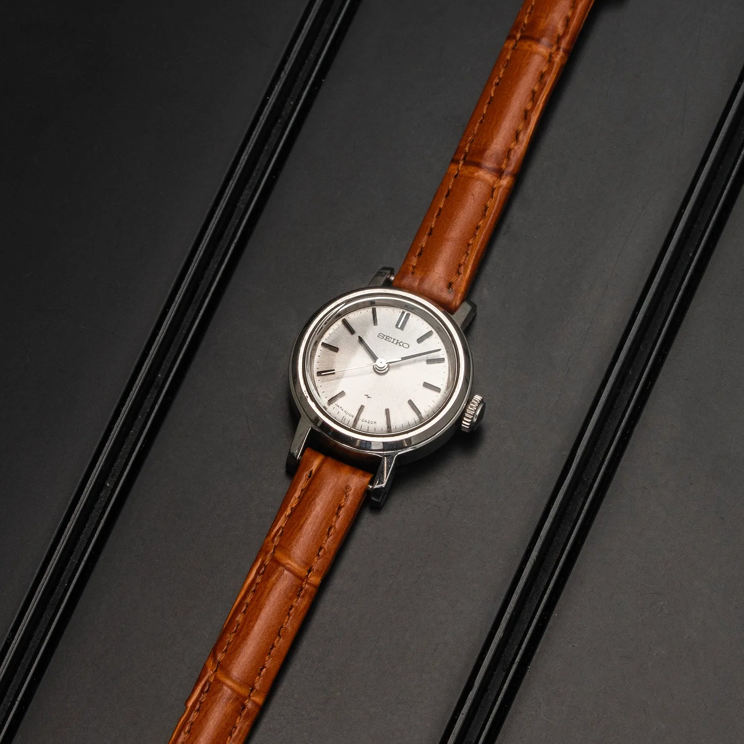 A vintage-style women's Seiko 1104-0150 wristwatch with a round silver-toned case, white dial, and brown leather strap, photographed diagonally on a dark gray background with black bars.