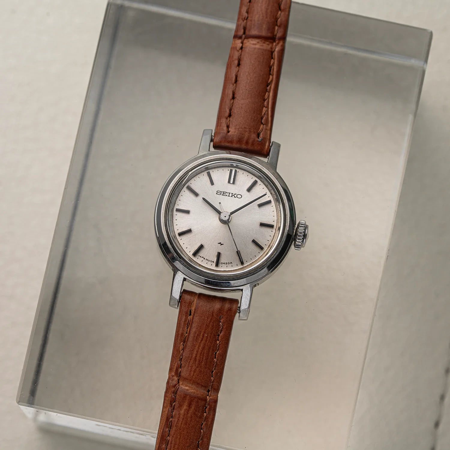 A vintage women's Seiko 1104-0150 wristwatch with a round silver-tone case, white dial, and brown leather strap, displayed on a clear acrylic stand against a light beige background.