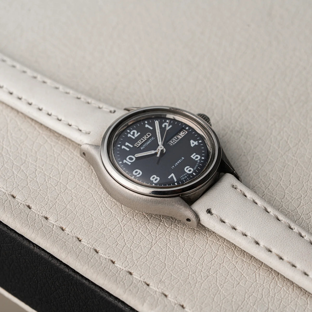 A Seiko, Ref. 2906-0230 wristwatch with a silver-toned metal case, black dial, and white leather strap, resting on a textured light beige leather surface.
