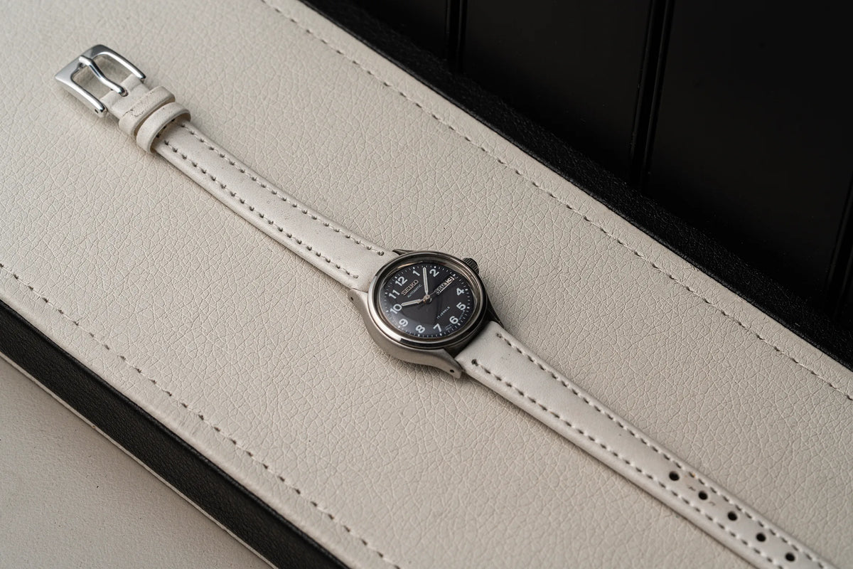 A minimalist wristwatch Seiko, Ref. 2906-0230 with a white leather strap and a round silver-tone case, resting diagonally on a textured cream leather surface with a black background.