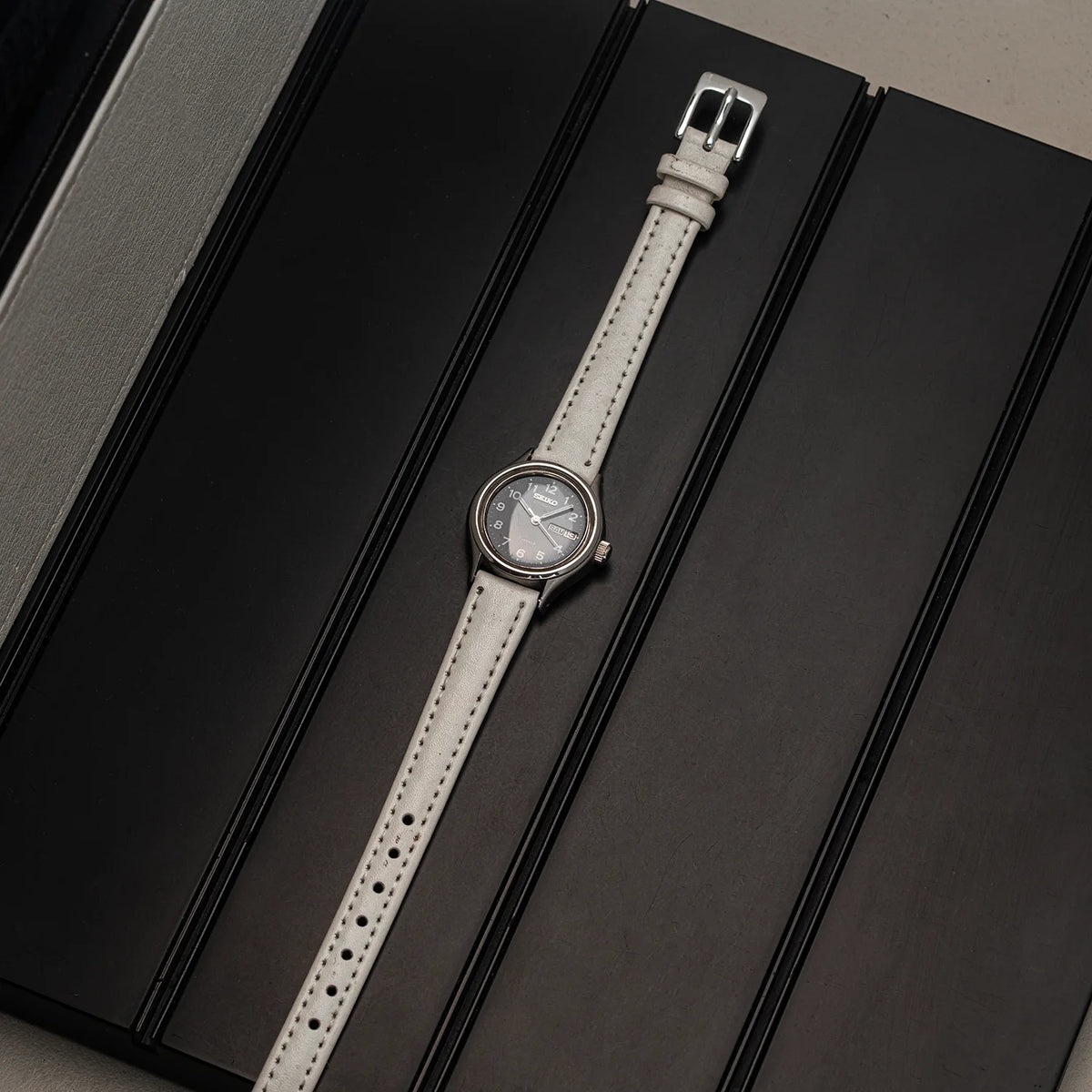 A wristwatch Seiko, Ref. 2906-0230 with a light gray leather strap and a silver-toned round case, displayed diagonally on a black textured surface.