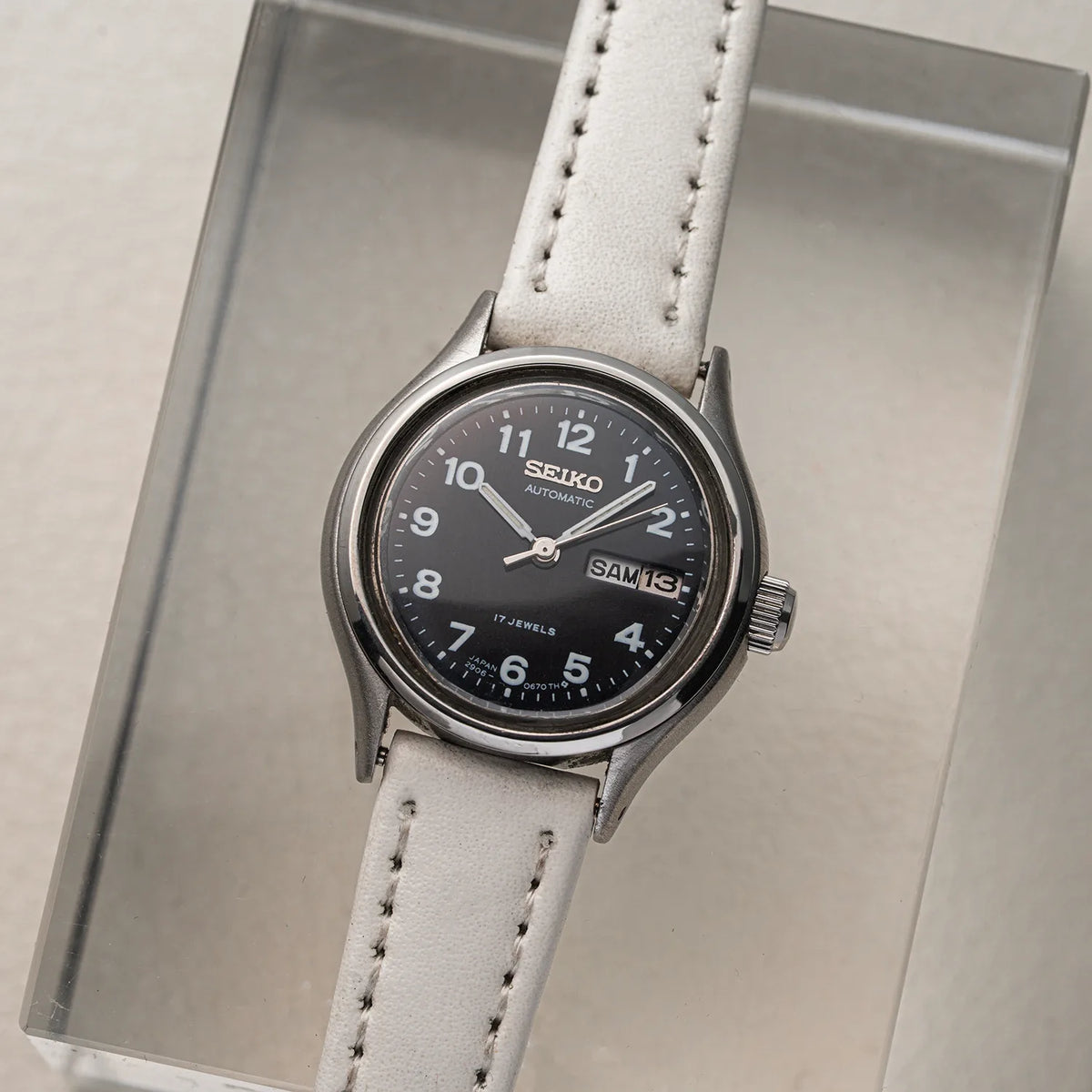 A Seiko, Ref. 2906-0230 women's watch with a black dial, silver-tone case, and a light beige leather strap, displayed on a clear acrylic stand.