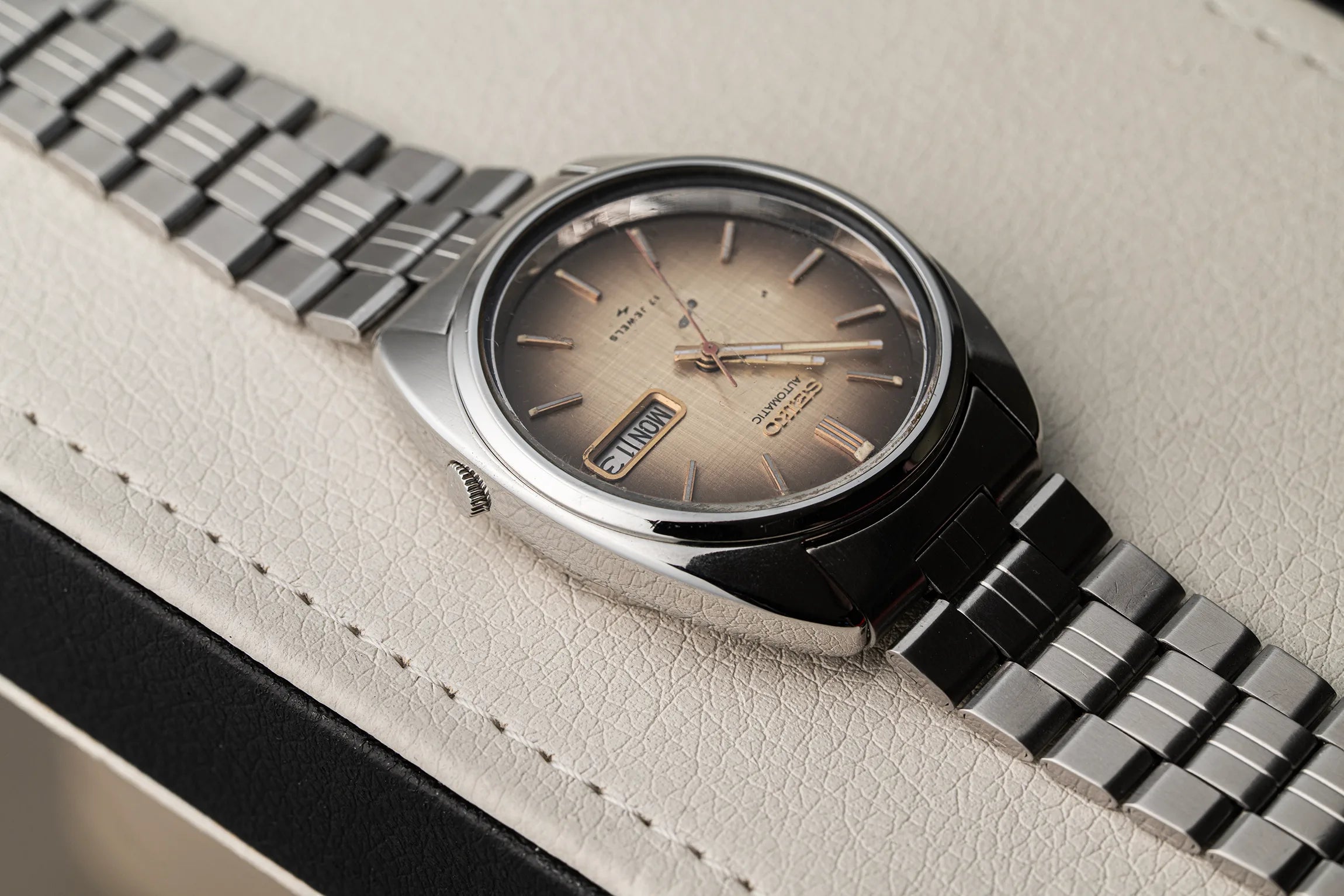 A close-up shot of a vintage-style wristwatch Seiko 7009-8081 with a silver metal link bracelet and a champagne-gold dial, resting on a textured light beige leather surface.