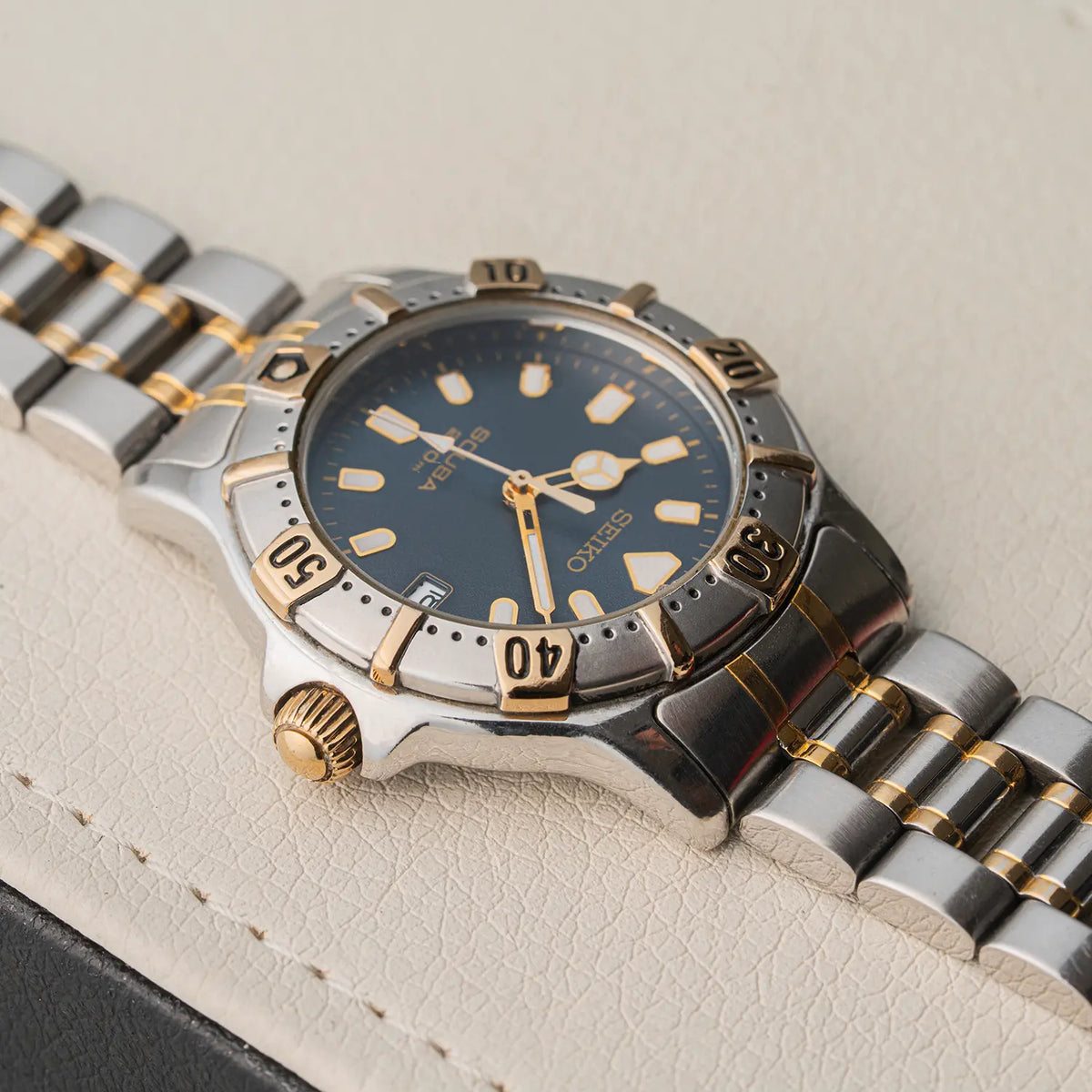 A close-up shot of a Seiko Scuba 200M, Ref. 7N35-6020, Cal. 7N35, Diving, 1992 watch with a two-tone silver and gold stainless steel bracelet, a blue dial, and a gold-tone crown, resting on a textured beige leather surface.