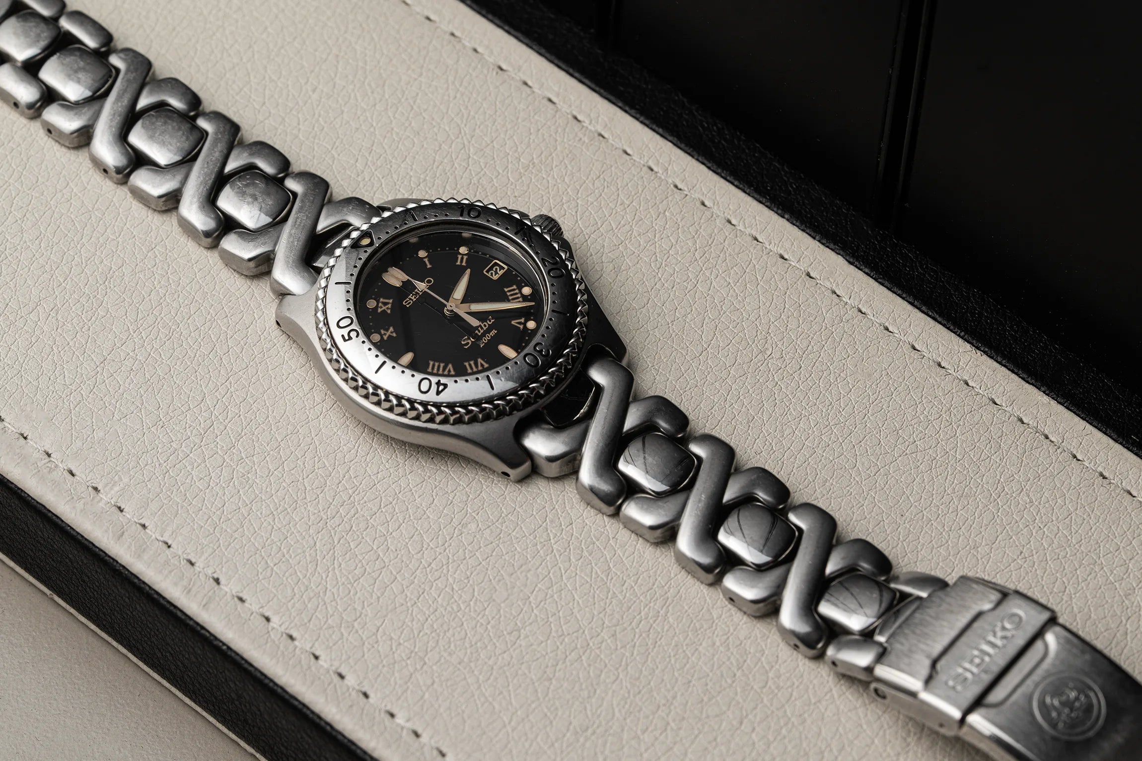 A close-up of a Seiko Scuba wristwatch with a silver-toned multi-link bracelet and a black dial, resting on a light beige leather surface.