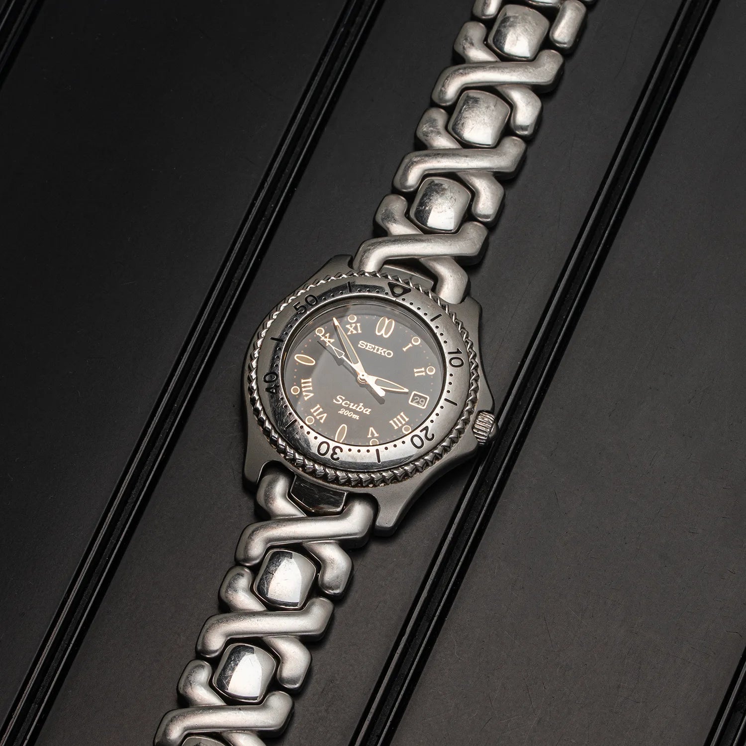 A Seiko Scuba stainless steel watch with a dark gray dial and a distinctive interlocking link bracelet, photographed diagonally on a dark textured background.