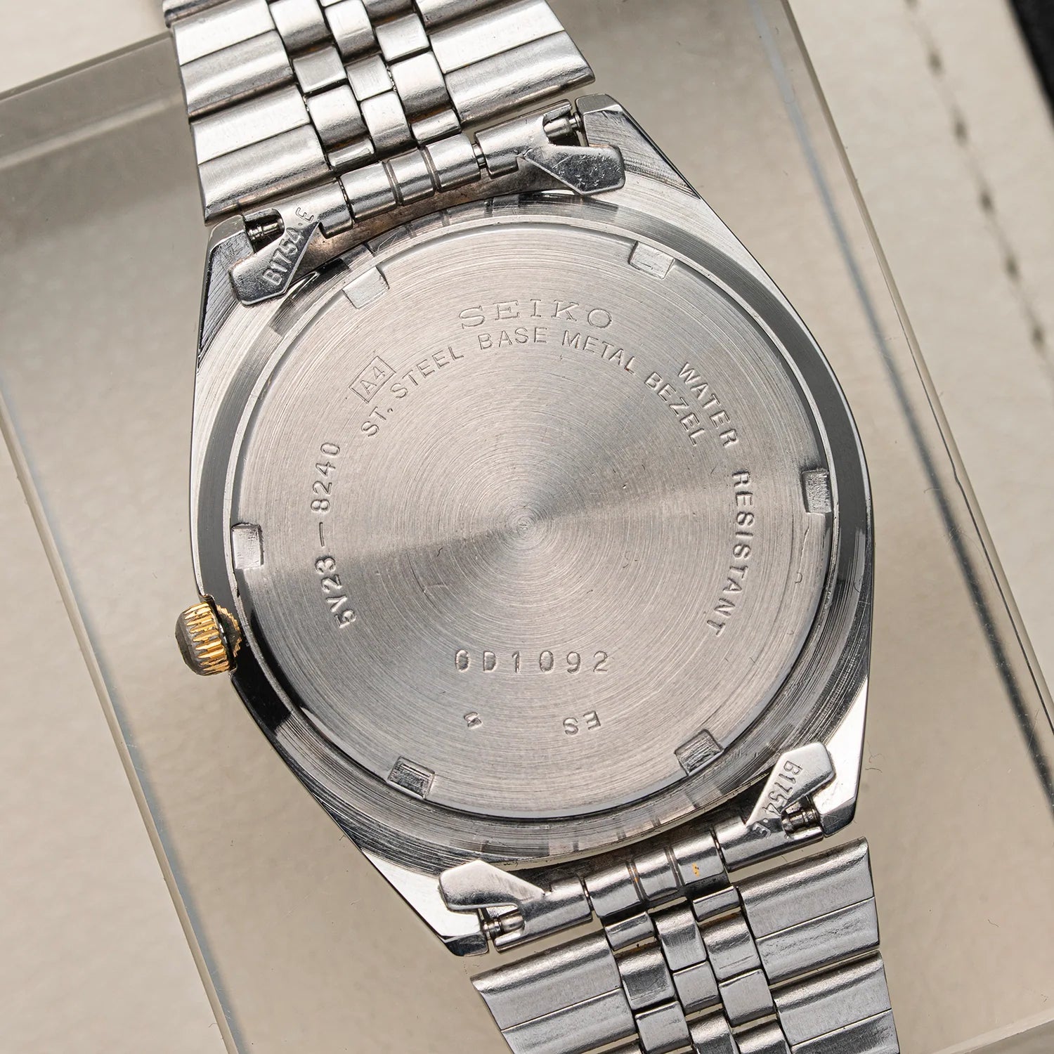 Close-up of the back of a silver stainless steel wristwatch Seiko SQ 5Y23-8240 with a metal link bracelet, resting on a light beige surface. The watch case and bracelet are polished silver with a small gold crown accent visible on the left side.