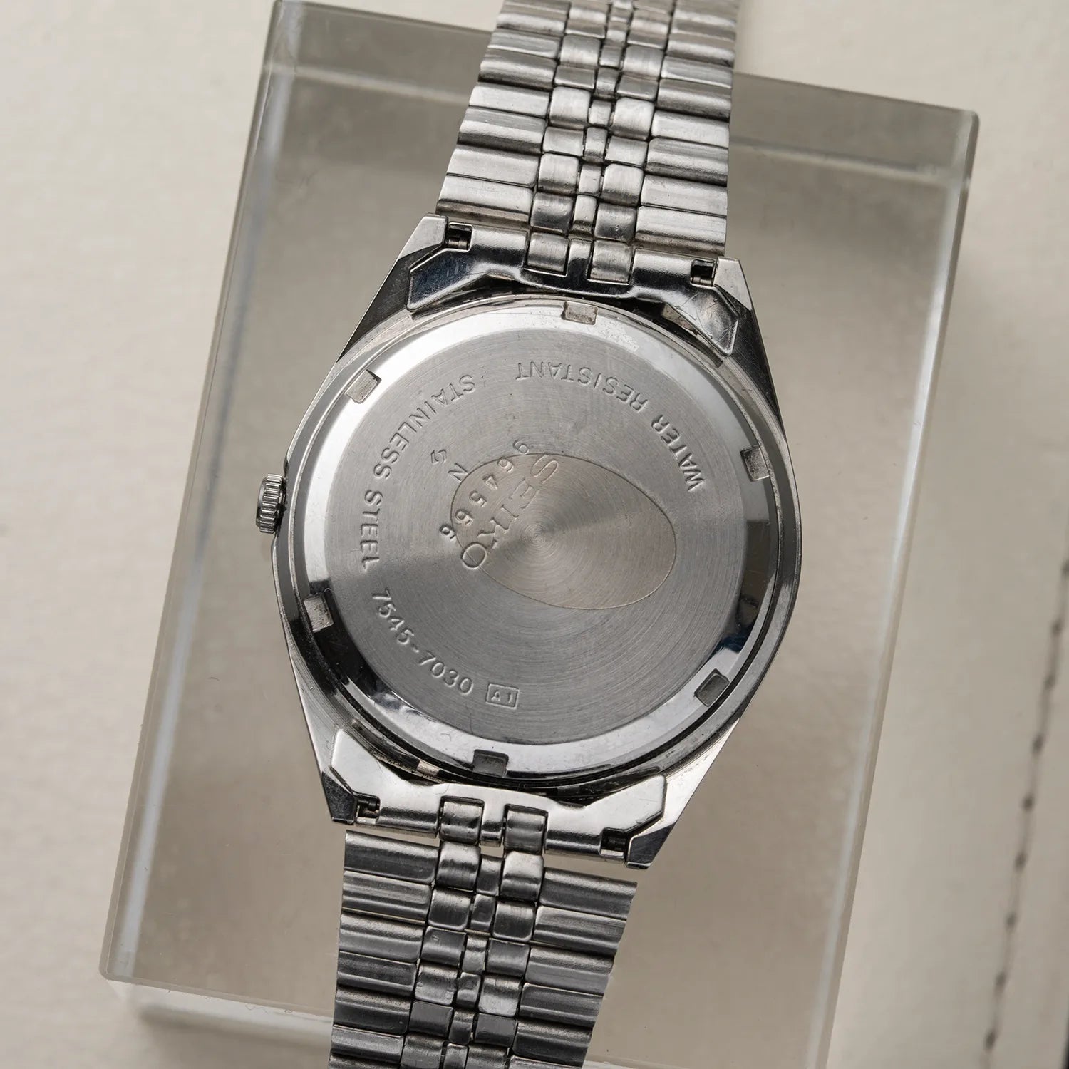 Silver stainless steel wristwatch Seiko SQ, Ref. 7545-7030 shown from the back, resting on a clear acrylic display block against a light beige background.