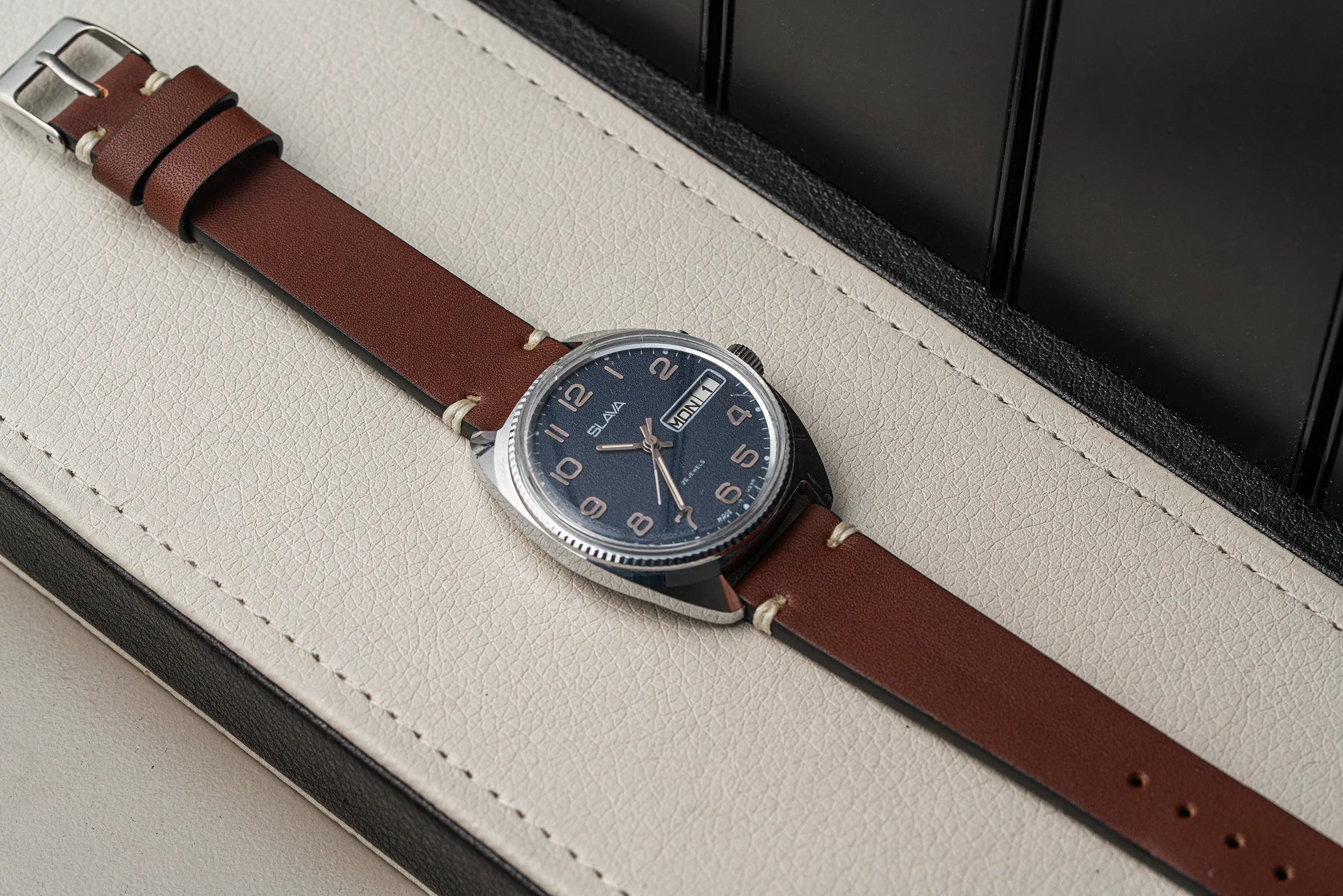 A wristwatch Slava Big Numerals with a brown leather strap and a dark blue dial, resting diagonally on a cream-colored textured surface.