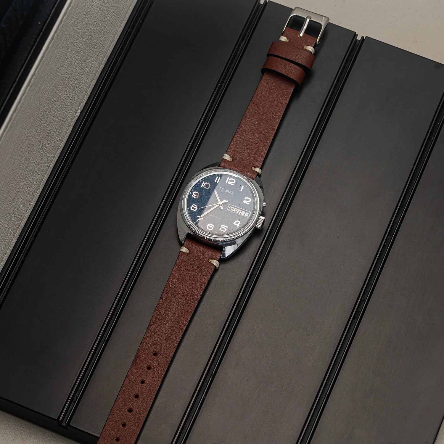 A wristwatch Slava Big Numerals with a brown leather strap and a dark blue dial, displayed diagonally on a black textured surface.