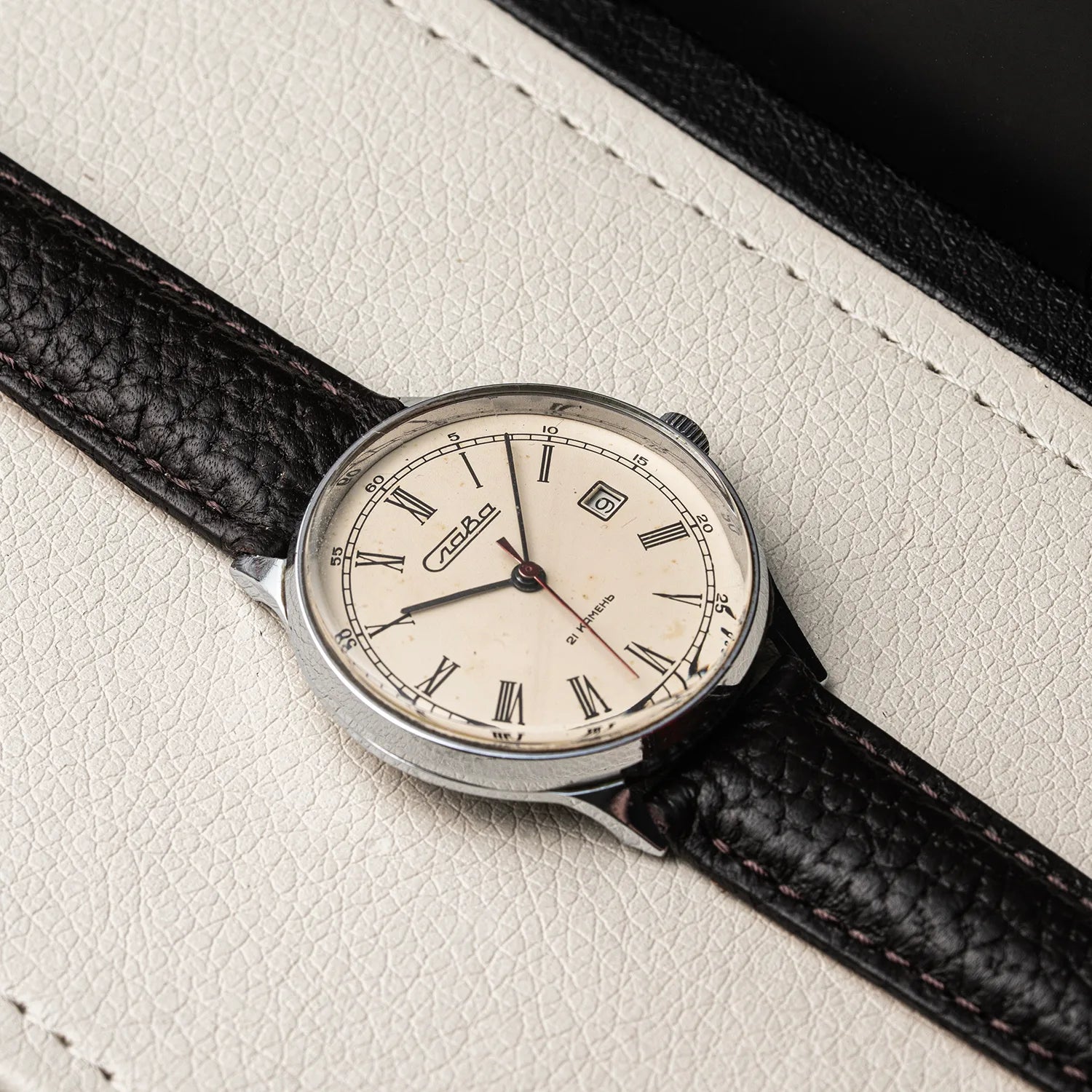 A vintage-style wristwatch Slava Roman Numerals with a silver-toned case, cream dial, and black textured leather strap, photographed diagonally on a light cream textured surface.